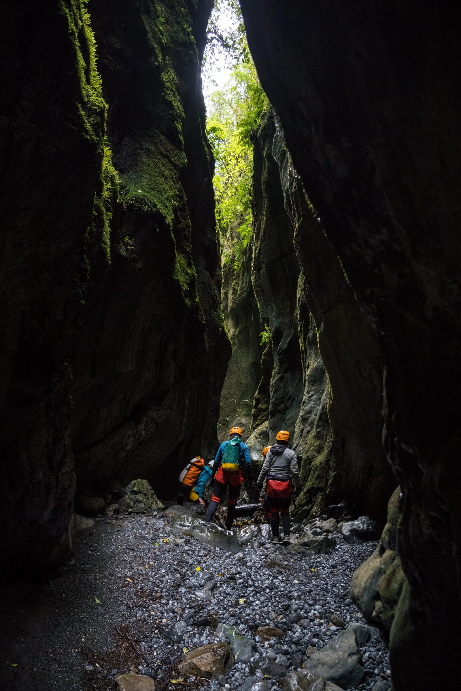 Group of adventurers wearing helmets and backpacks exploring a narrow, dark canyon with moss-covered rock walls and a shallow stream.