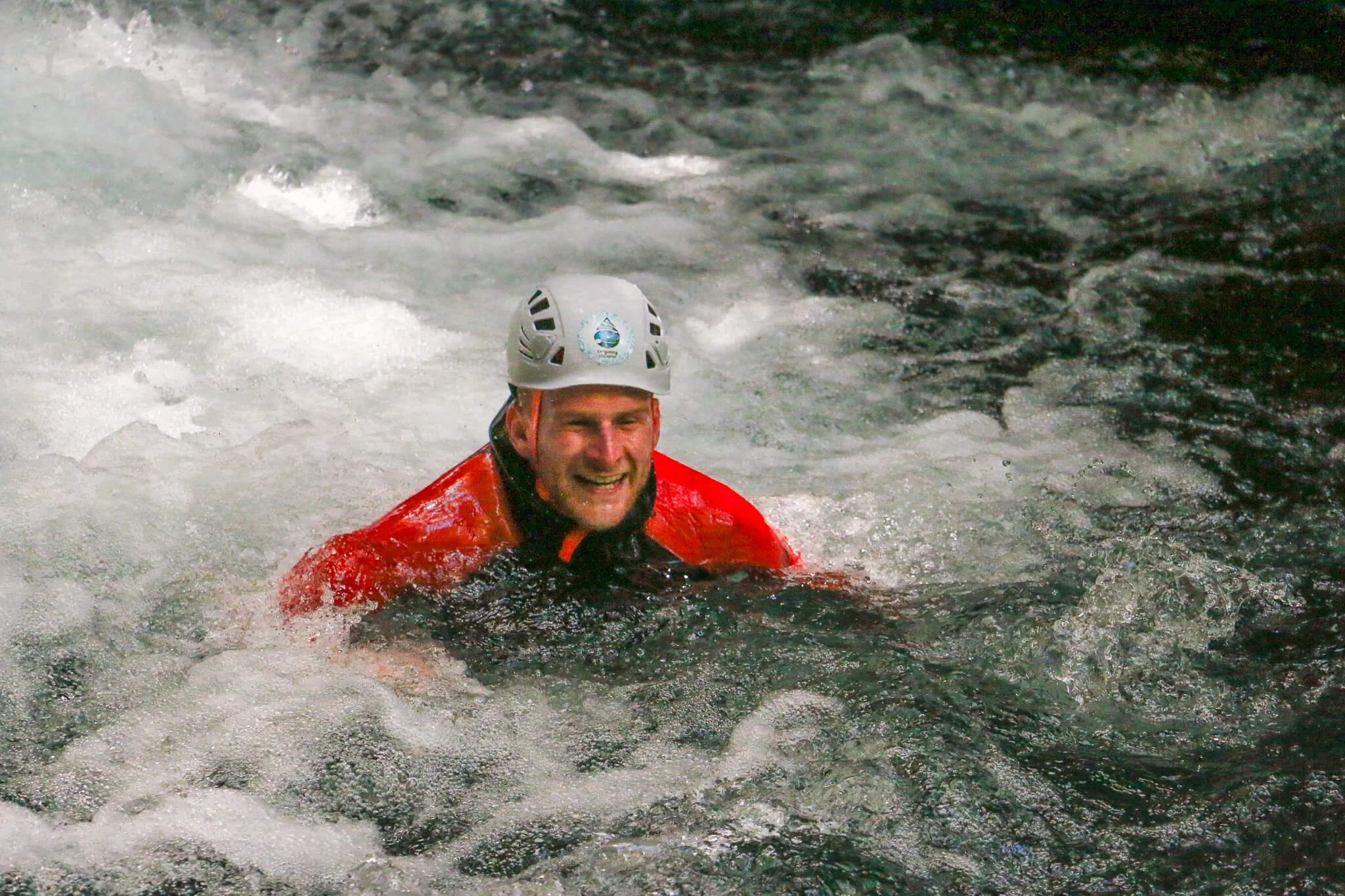 A man wearing a helmet and red jacket smiling while swimming in a turbulent river with white water rapids.