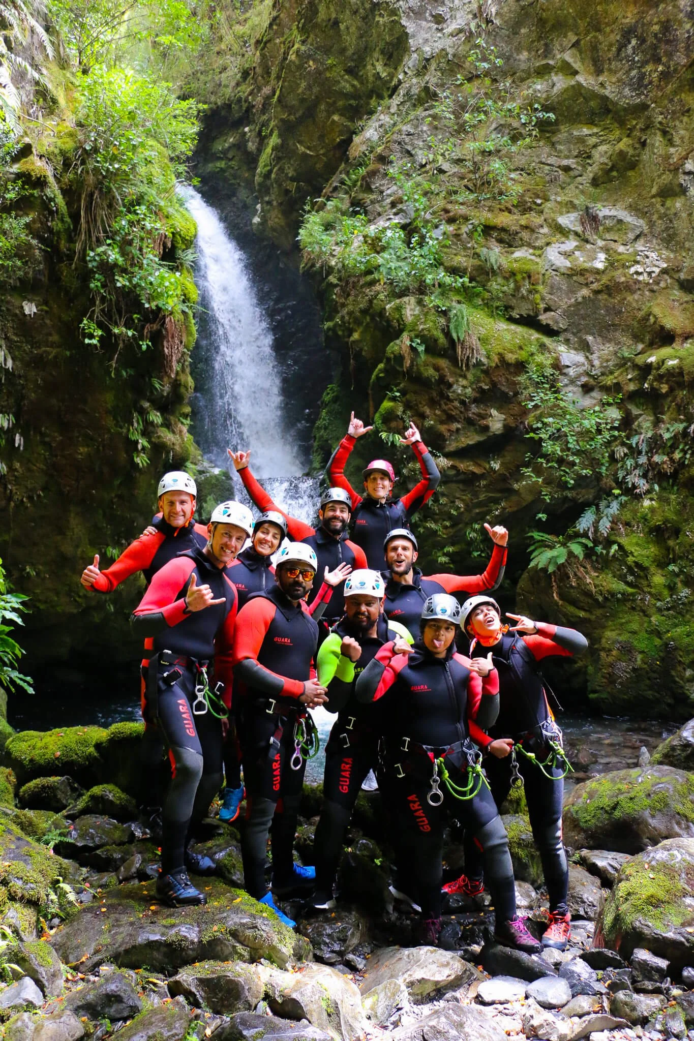 A group of people dressed in black and red protective gear standing on rocks in front of a waterfall and lush green foliage in a forest.