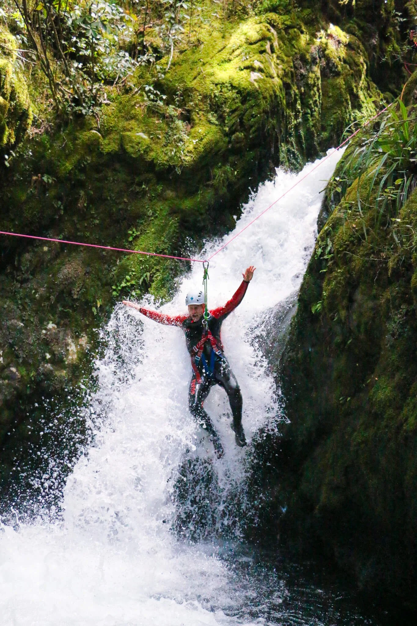 Person wearing helmet and wetsuit sliding down a waterfall on a zip line in a lush, moss-covered canyon surrounded by green rocks and vegetation.