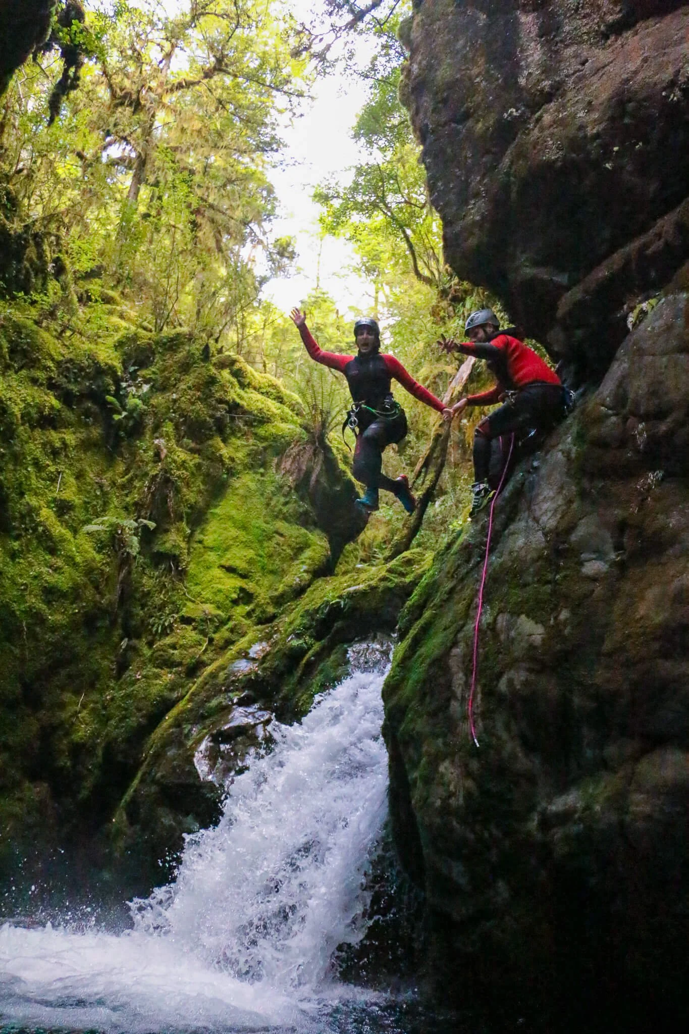 Two people are canyoning in a narrow, moss-covered canyon with rushing water. They are wearing helmets and wetsuits, holding hands, and one is waving at the camera, surrounded by lush green foliage.