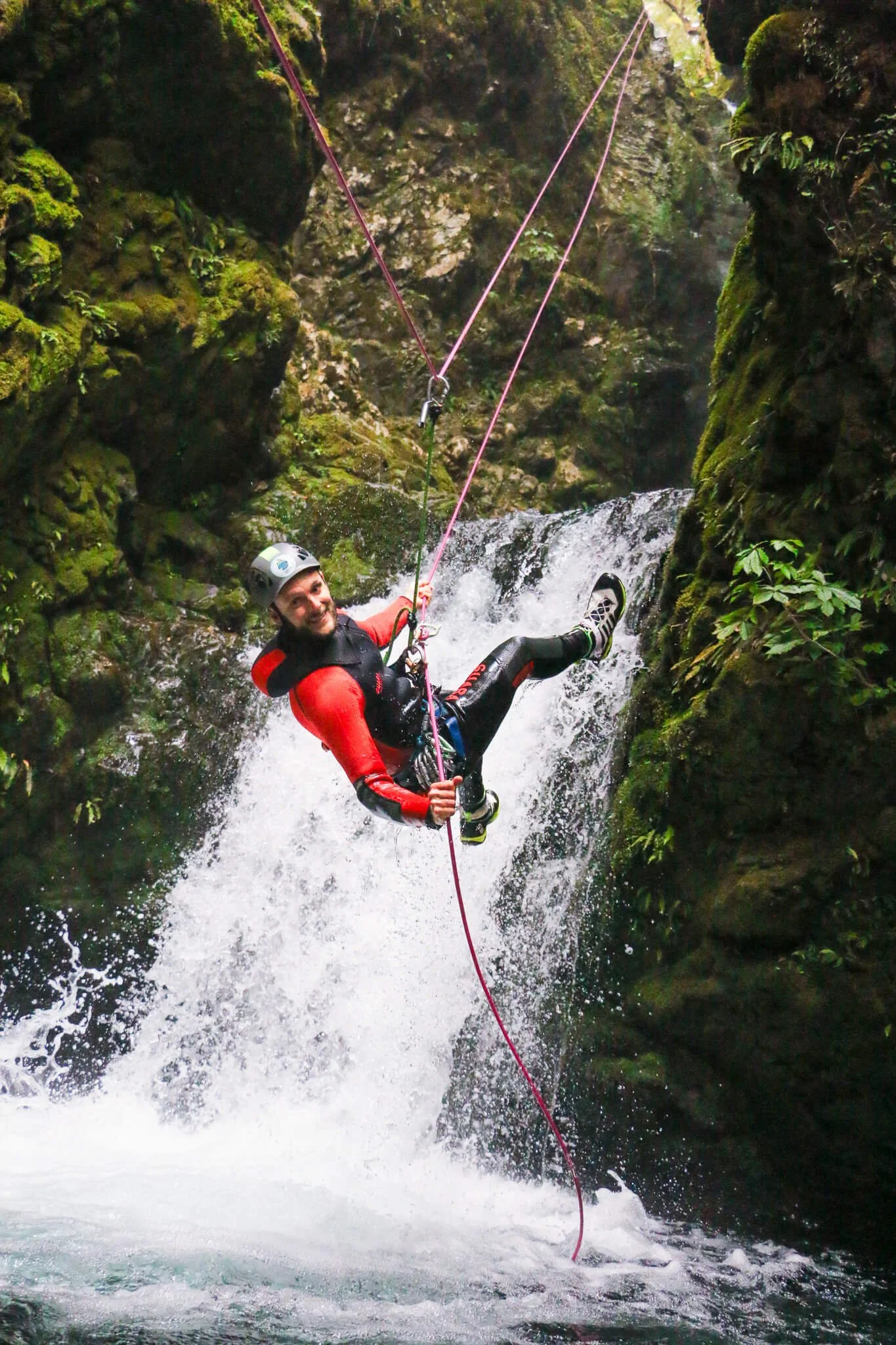 A person wearing a helmet and harness is rappelling down a moss-covered rocky canyon wall over a rushing stream, holding a red rope for control.