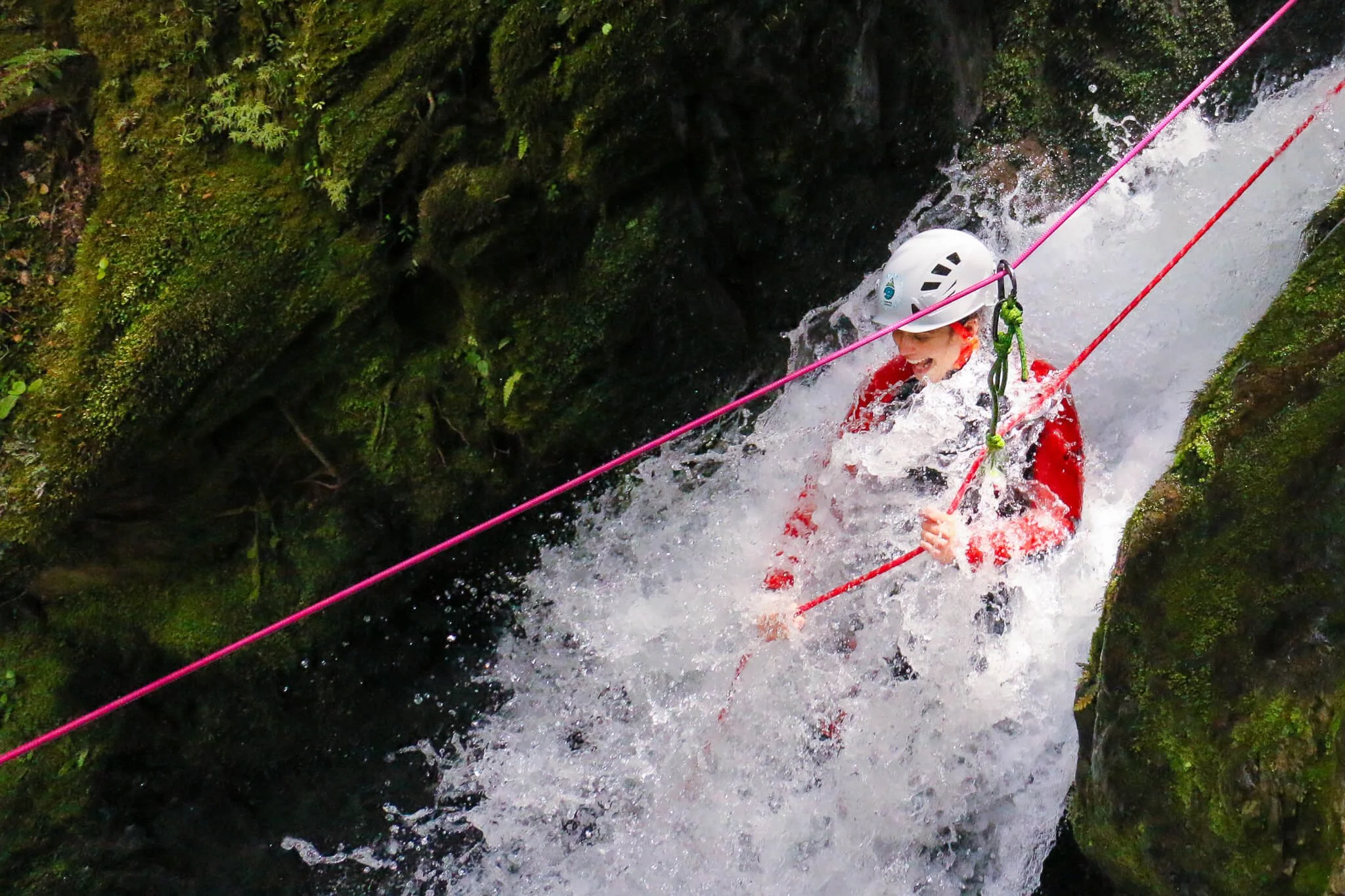 A person wearing a white helmet and red jacket is enjoying white water rafting, gripping the safety rope while navigating through a narrow section of a moss-covered rocky stream.