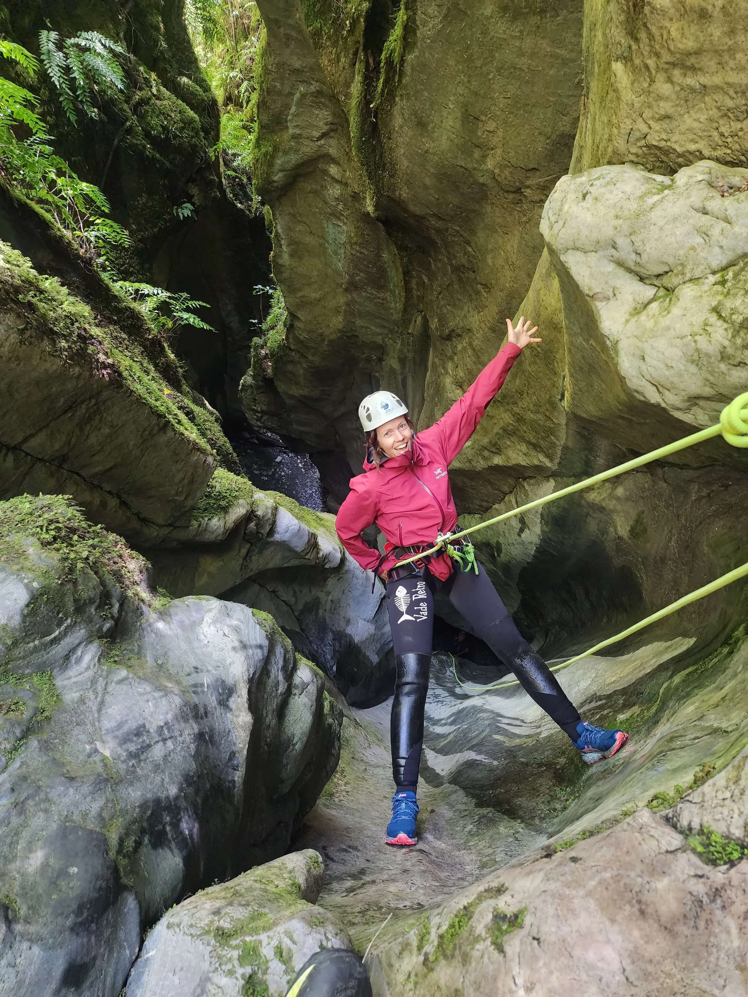 A person in outdoor climbing gear, including a helmet and waterproof clothing, smiling and standing in a narrow rocky canyon with moss-covered rocks and a rope.