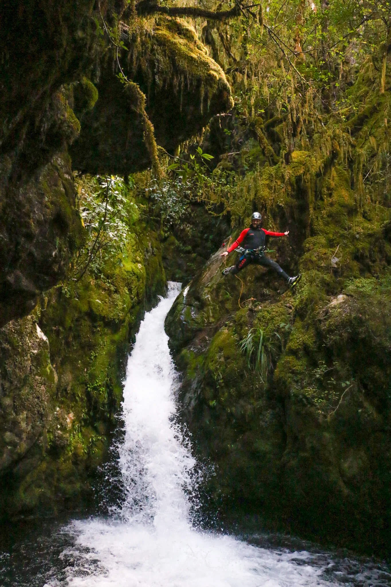 A person in outdoor gear and a helmet rock climbing on moss-covered rocks beside a rushing mountain stream surrounded by lush greenery.