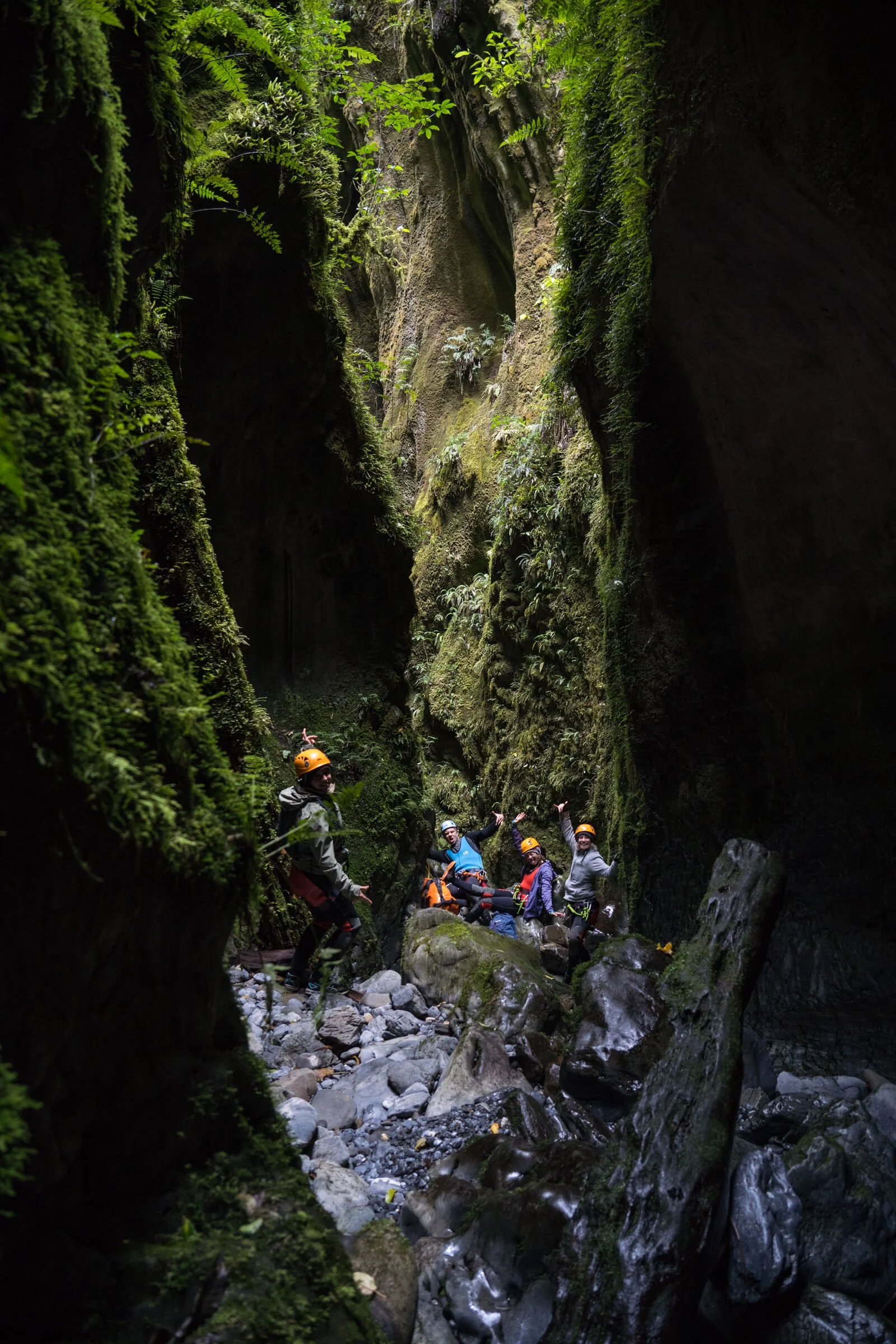 Six people in helmets and outdoor gear sitting and standing on rocks inside a narrow canyon with moss-covered walls and a small stream.