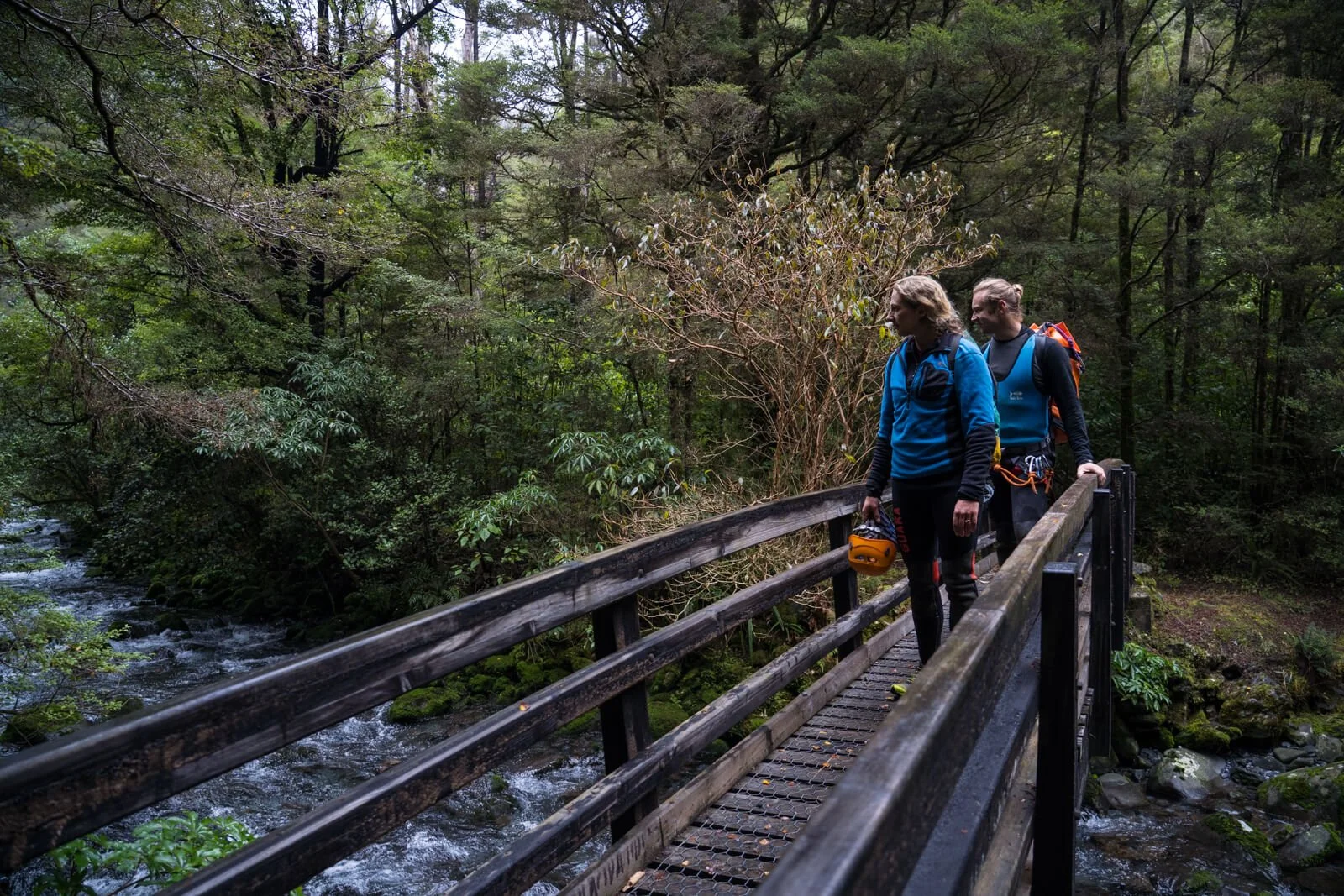 Two women walking across a wooden bridge over a stream in a lush green forest, wearing outdoor hiking gear and carrying helmets.