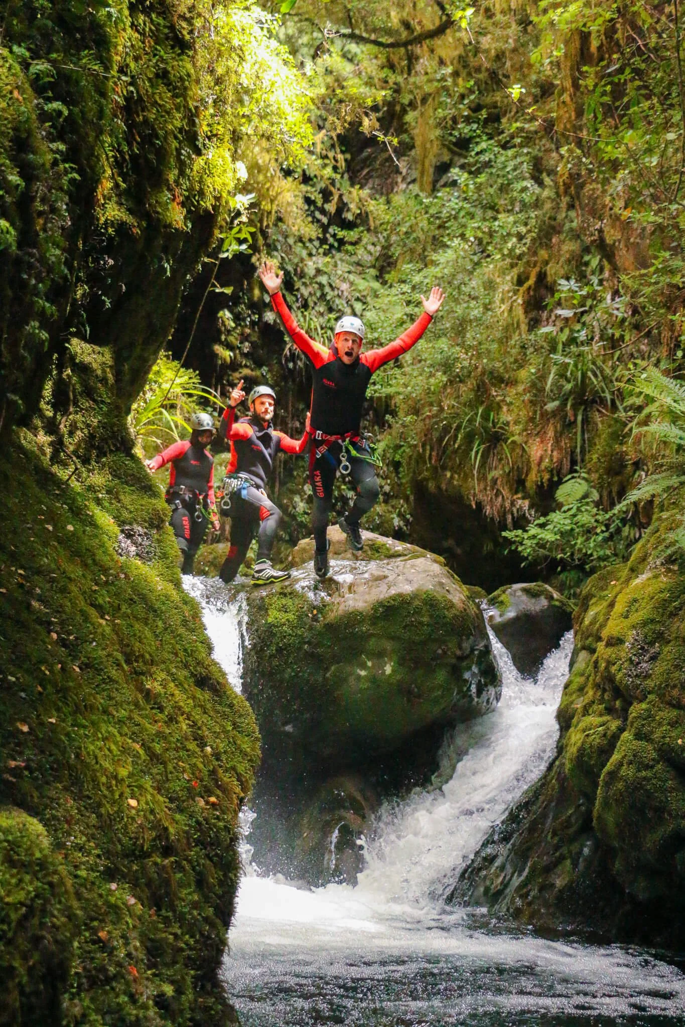 Three adventurers in safety gear, including helmets and wetsuits, hike through a lush, mossy canyon with a flowing stream, surrounded by dense green foliage.
