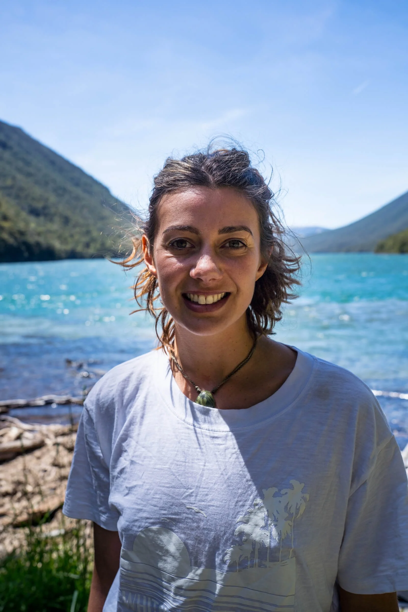 Camille smiling outdoors near a body of water, with hills or mountains in the background under a clear blue sky.