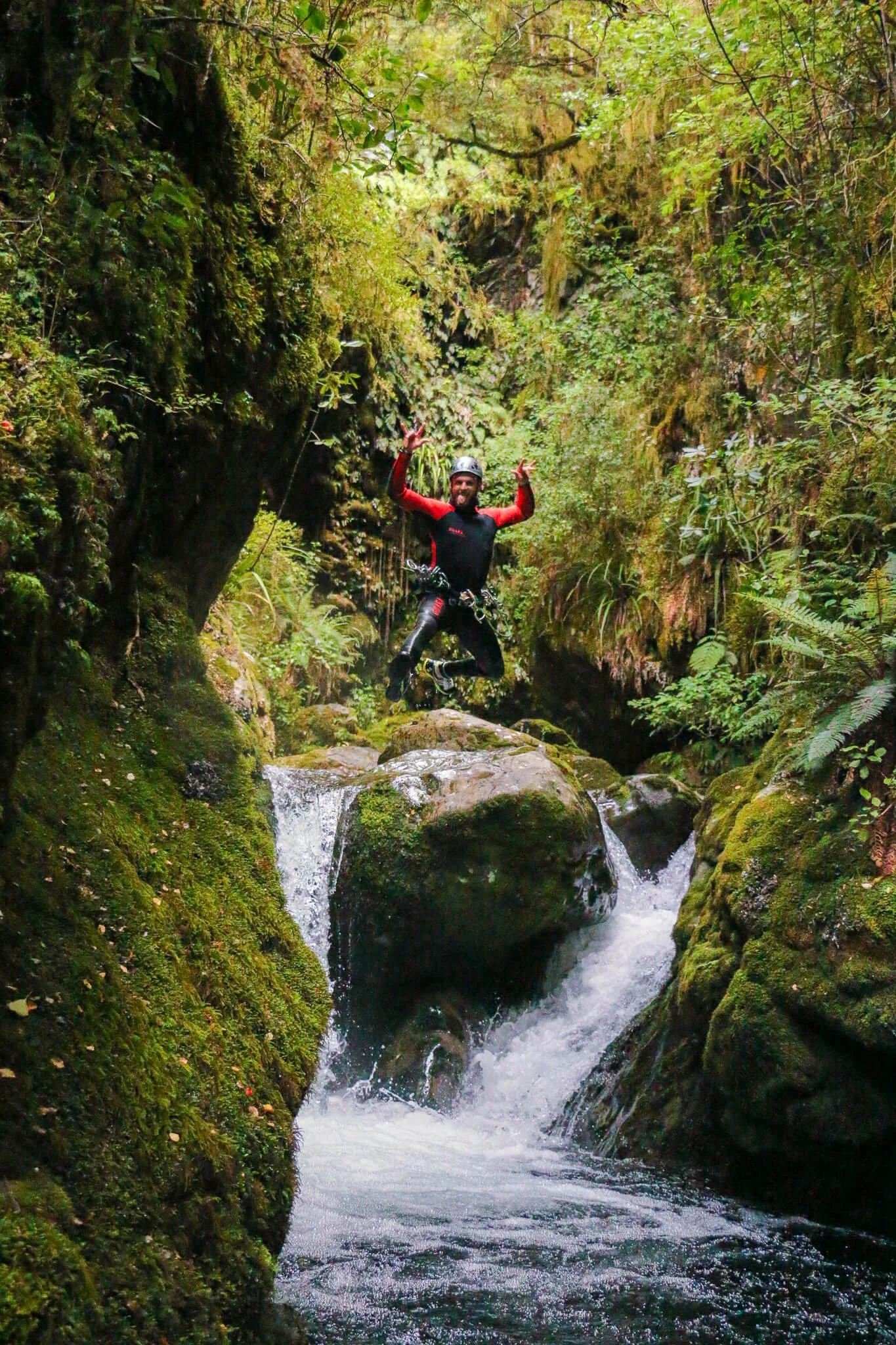 A person wearing a helmet and waterproof clothing is jumping across rocks over a rushing stream in a lush, green forest.