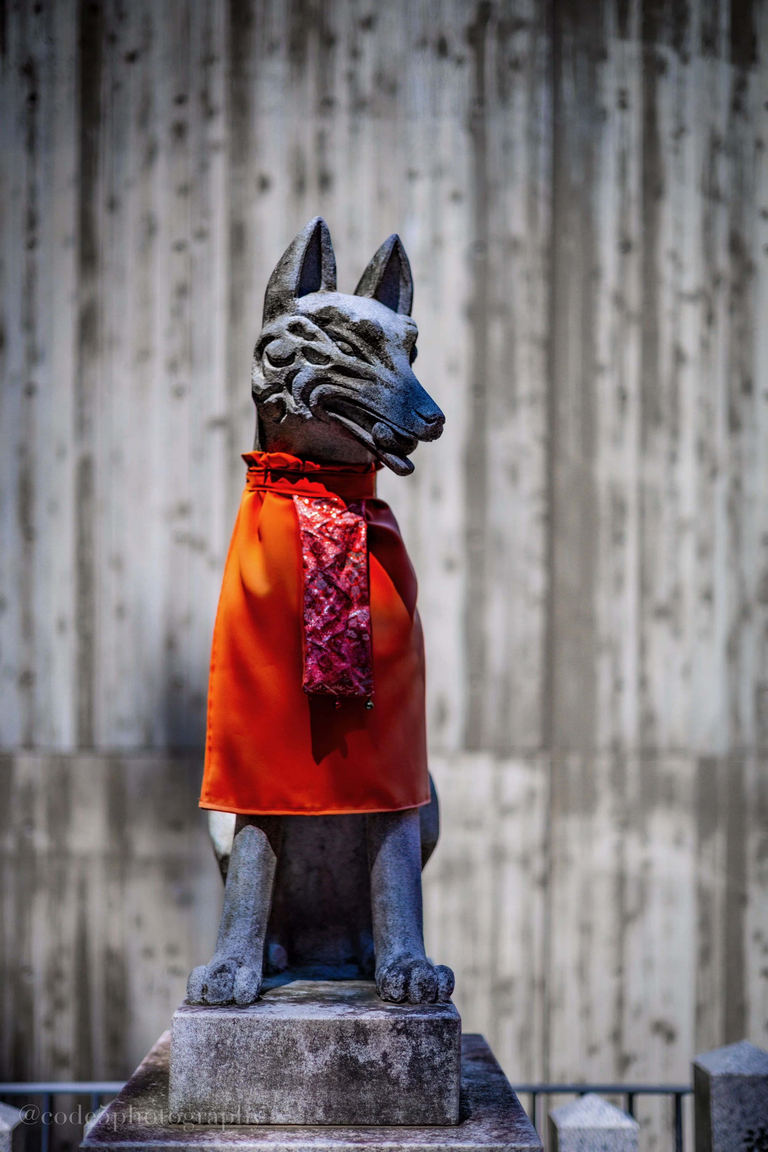A stone statue of a dog, possibly a German Shepherd, wearing an orange cape with red and gold trim, standing on a stone base with a gray, weathered wooden fence background.