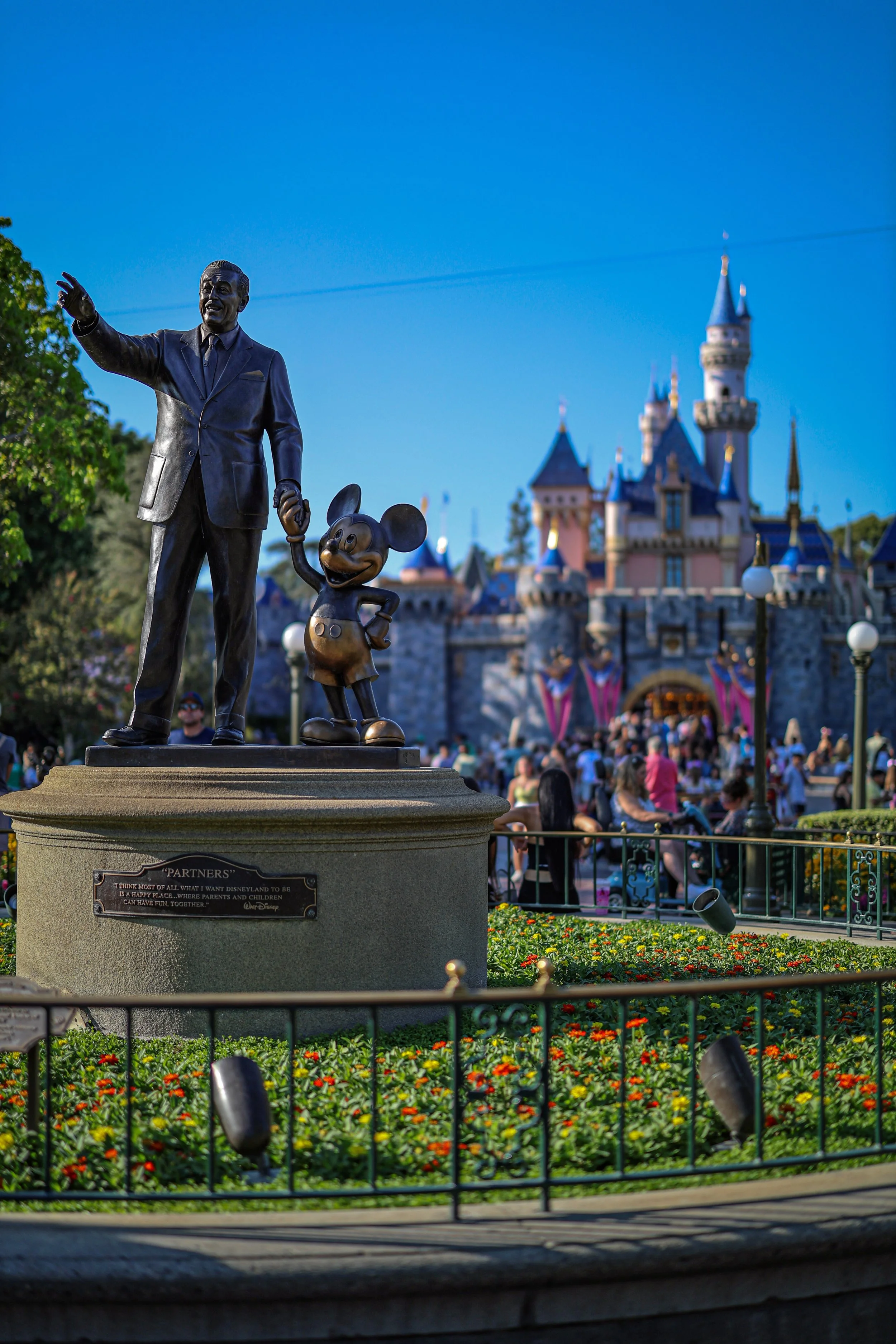 The statue of Walt Disney and Mickey Mouse in front of Sleeping Beauty Castle at Disneyland during the daytime, with visitors and colorful flowers in the background.