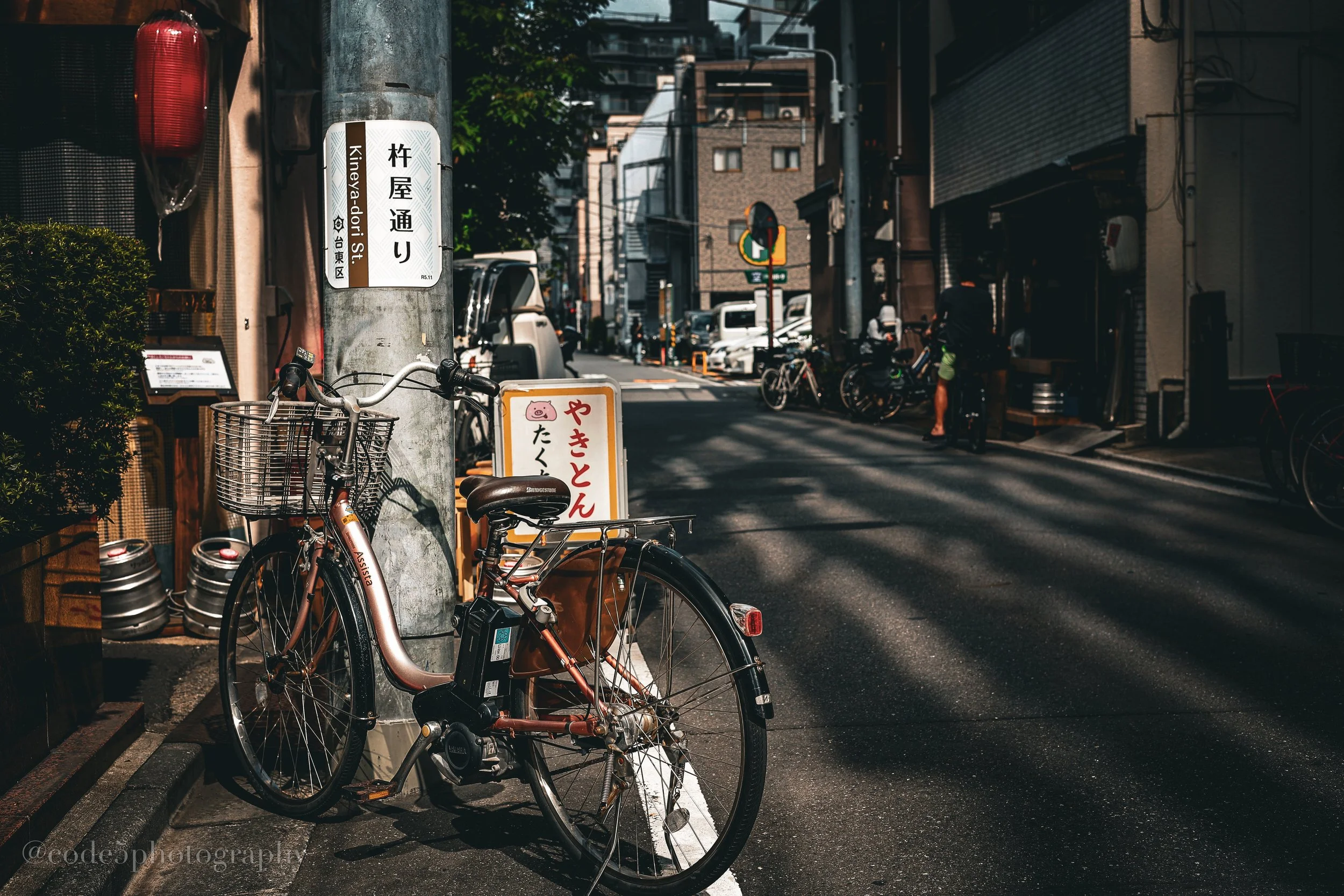 A bicycle parked on the sidewalk next to a utility pole with Japanese street signs, in a narrow urban street with buildings and parked cars, shot in bright daylight.