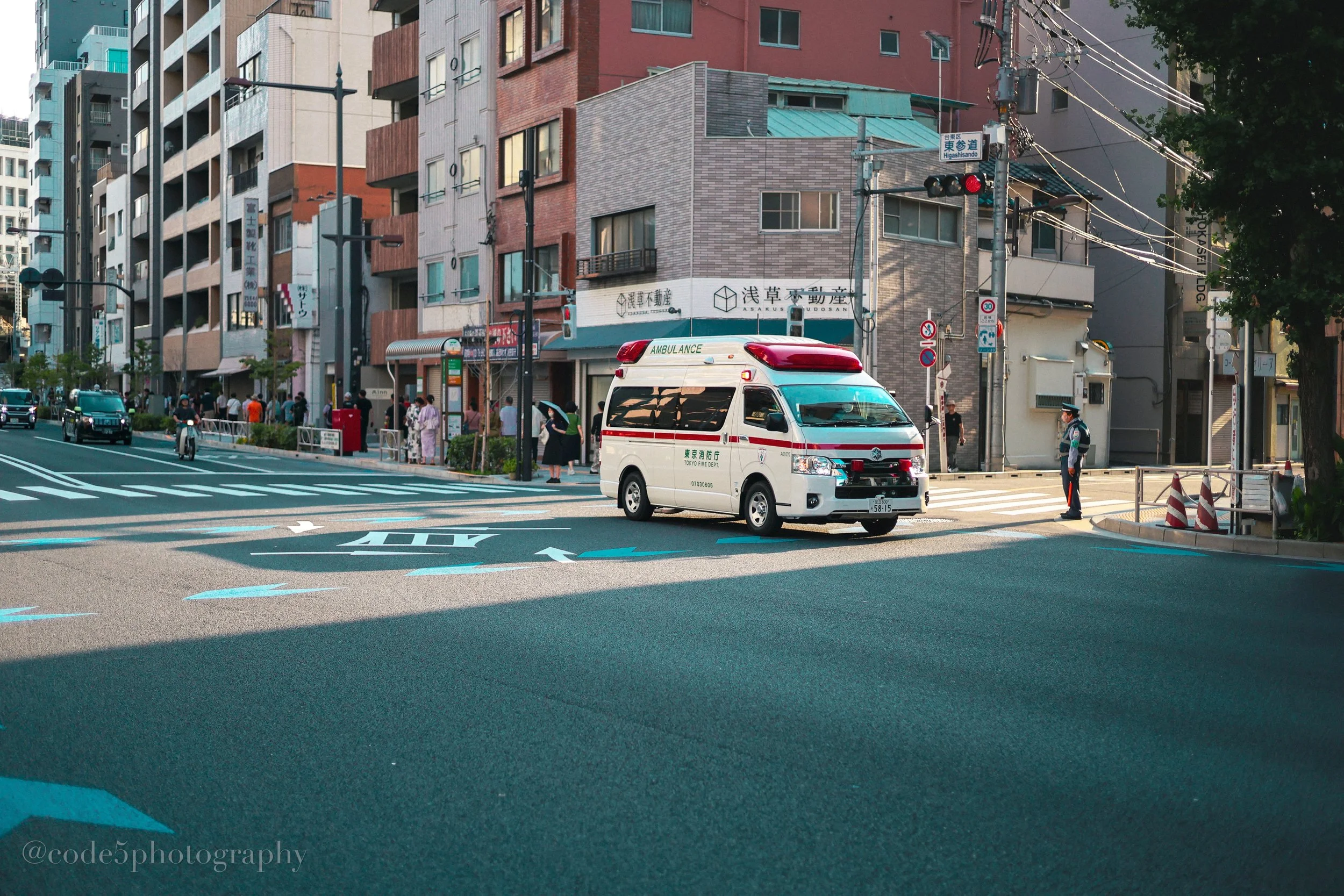 An urban street scene with a white ambulance vehicle stopped at a crosswalk, surrounded by pedestrians and cars, in front of multistory buildings with various signs and traffic signals.