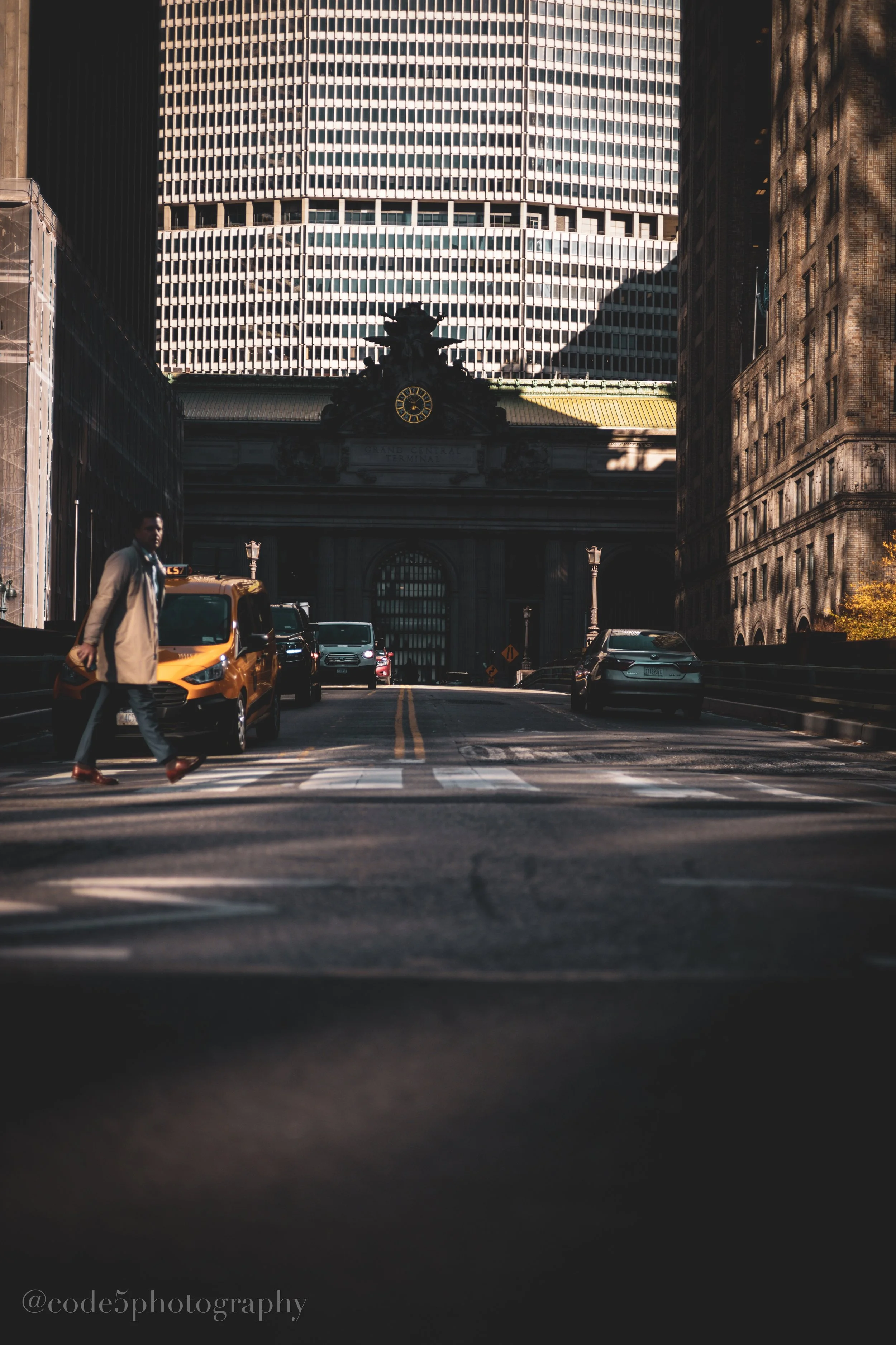 A city street scene with tall buildings on either side, a person crossing the street, and a yellow taxi car in the foreground. The buildings are modern, with one imposing skyscraper with a grid-like facade, and an older building with detailed archite