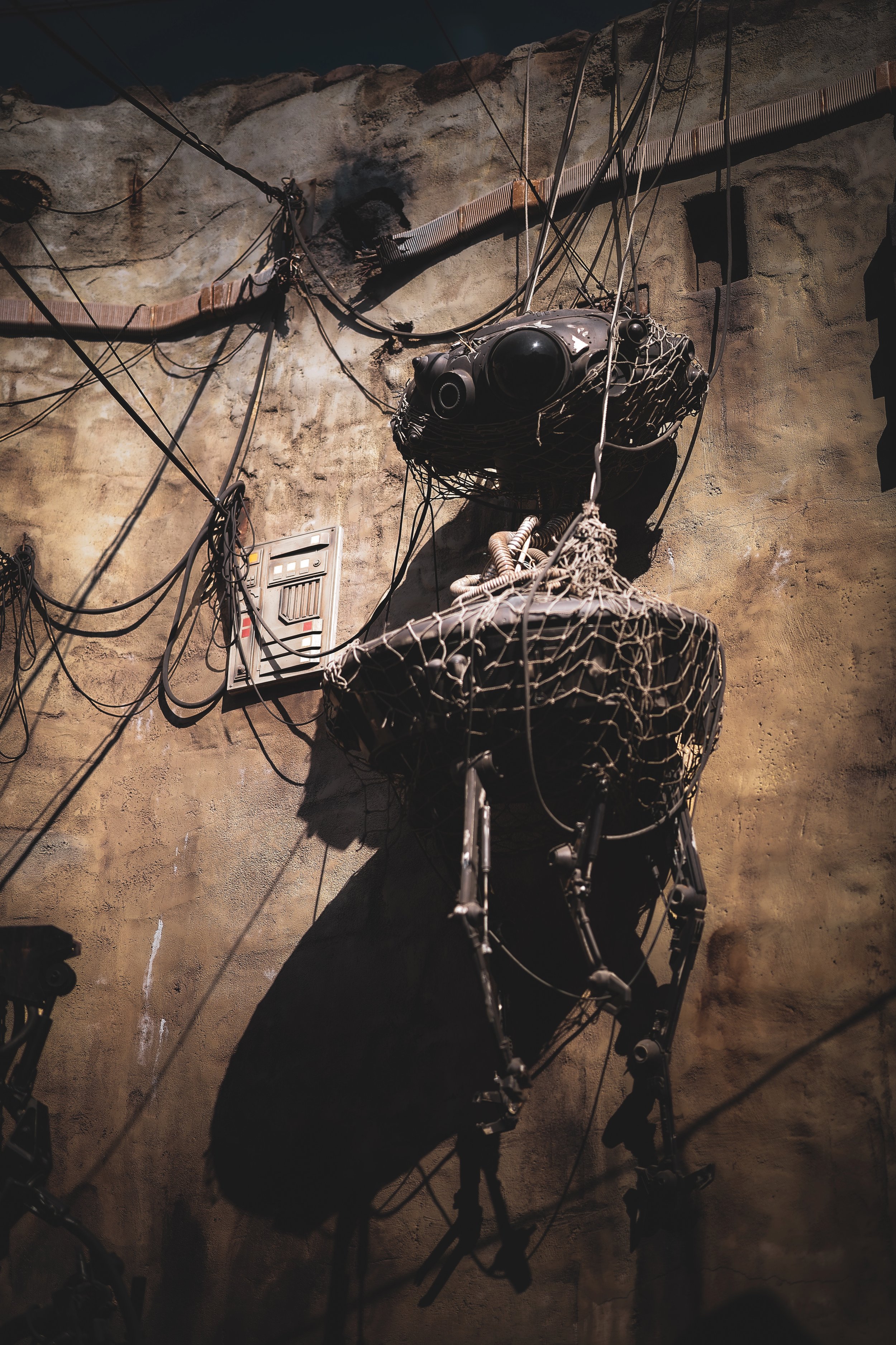 Sculpture of a human figure made from black material with wires and mechanical parts, set against a textured, weathered wall with stored wires and electrical panels.