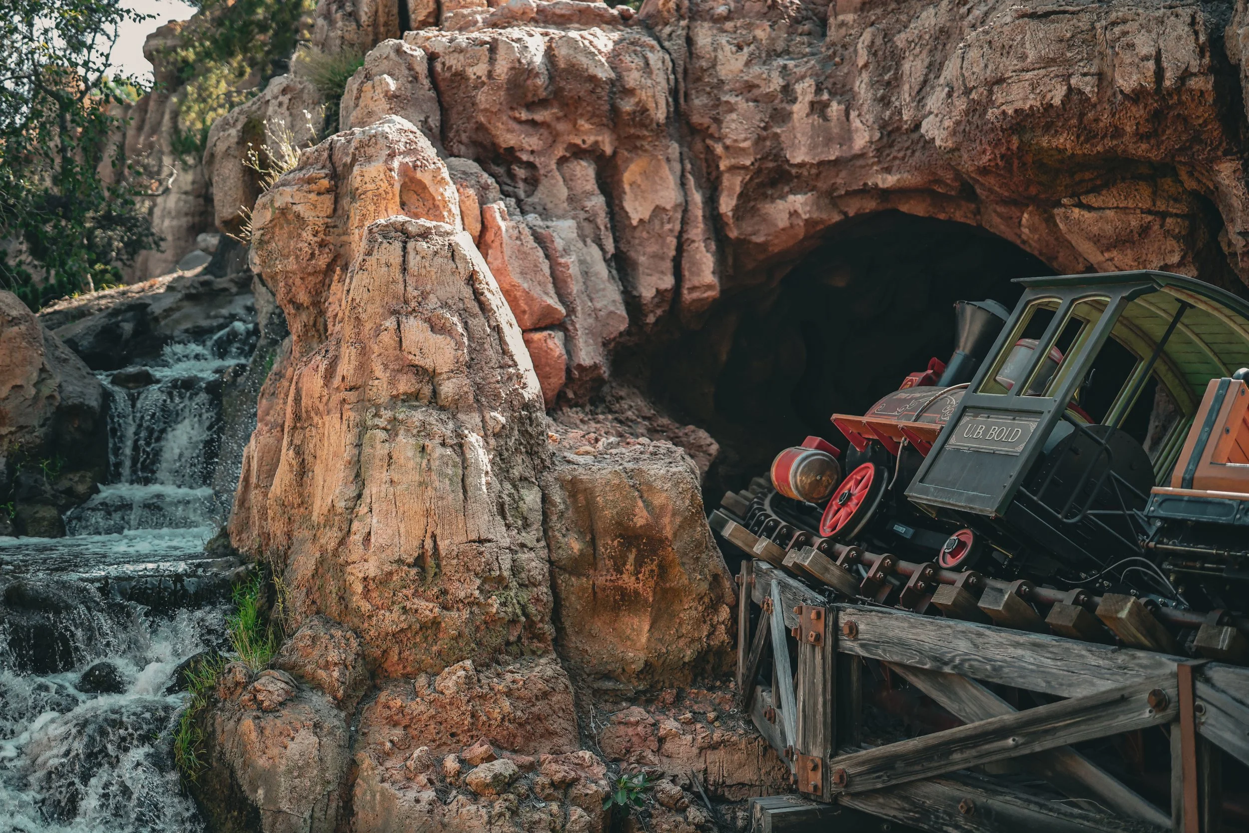 A small train with a vintage design on a wooden track, positioned near a cave entrance surrounded by rocky terrain and a flowing stream.