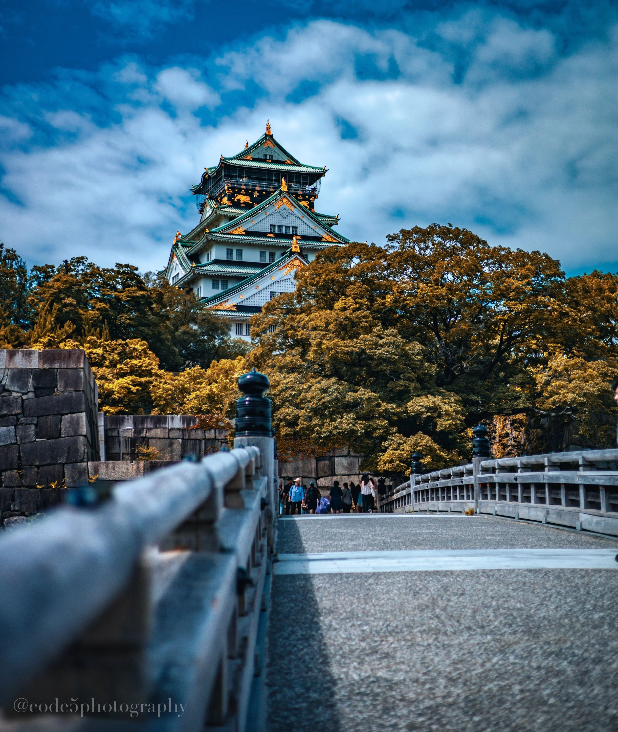 A traditional Japanese castle with green roofs and golden accents, surrounded by trees with autumn foliage, viewed from a low angle with a bridge in the foreground and a sky filled with clouds.