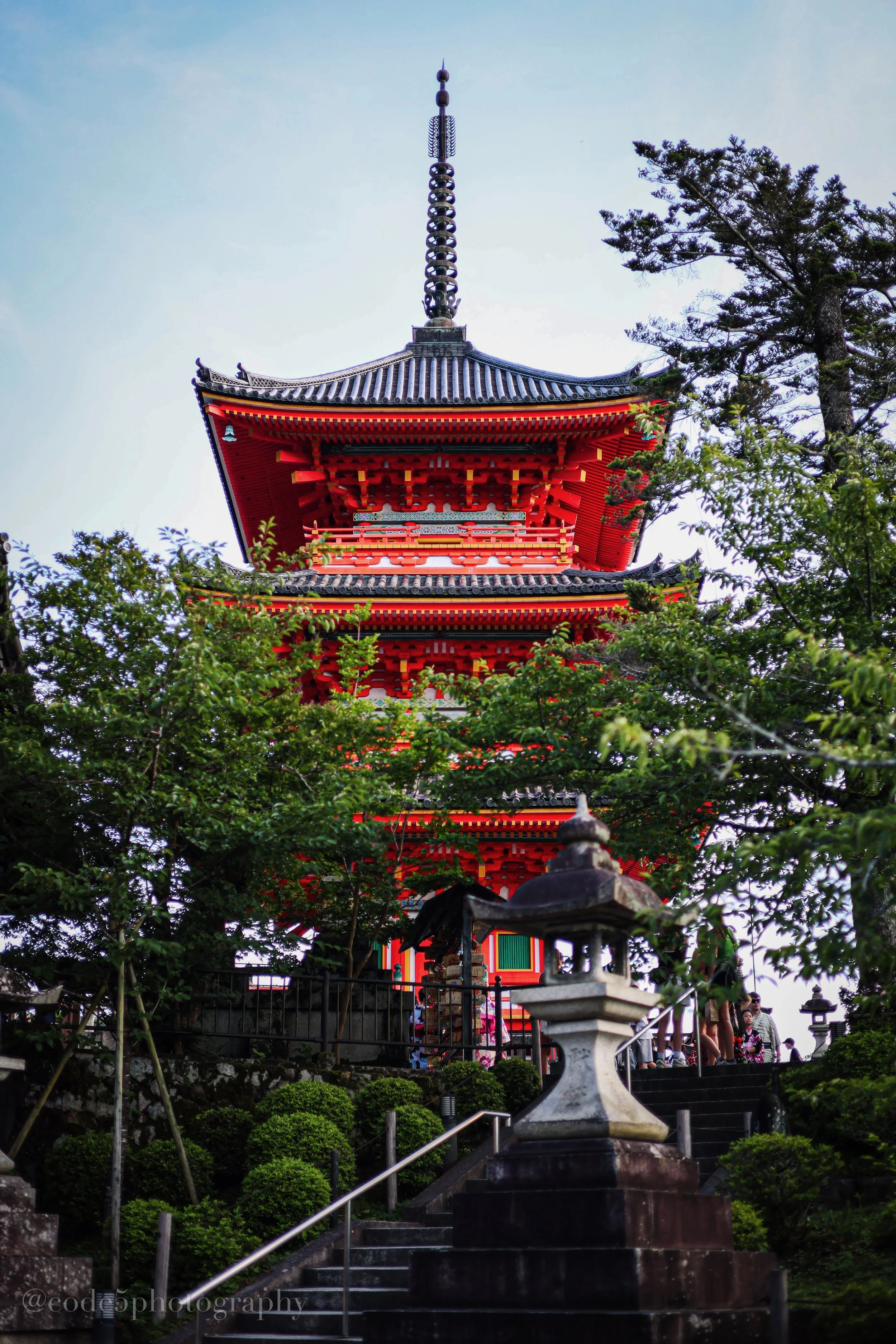 A traditional Japanese pagoda surrounded by greenery with a stone lantern in front, framed by trees under a partly cloudy sky.