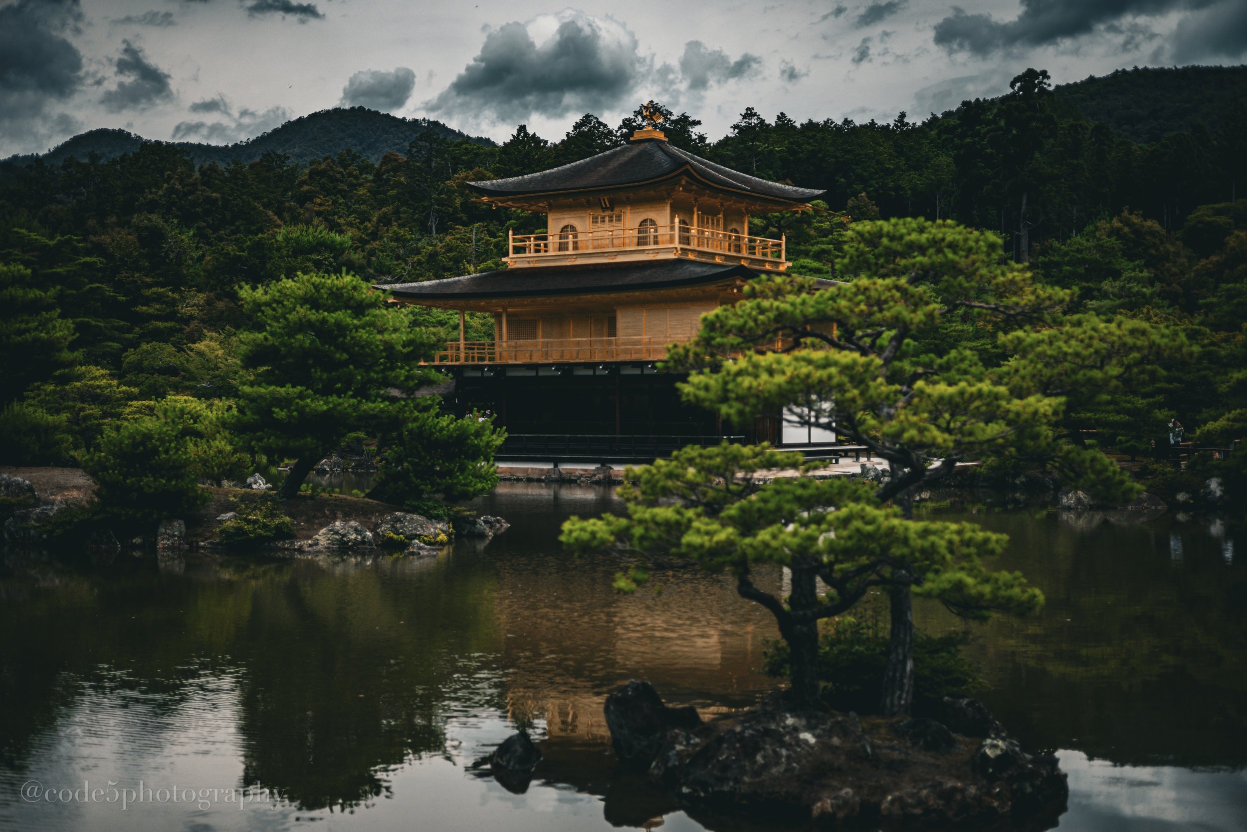 Traditional Japanese pagoda surrounded by lush greenery and trees, with a body of water in the foreground and mountains and cloudy sky in the background.