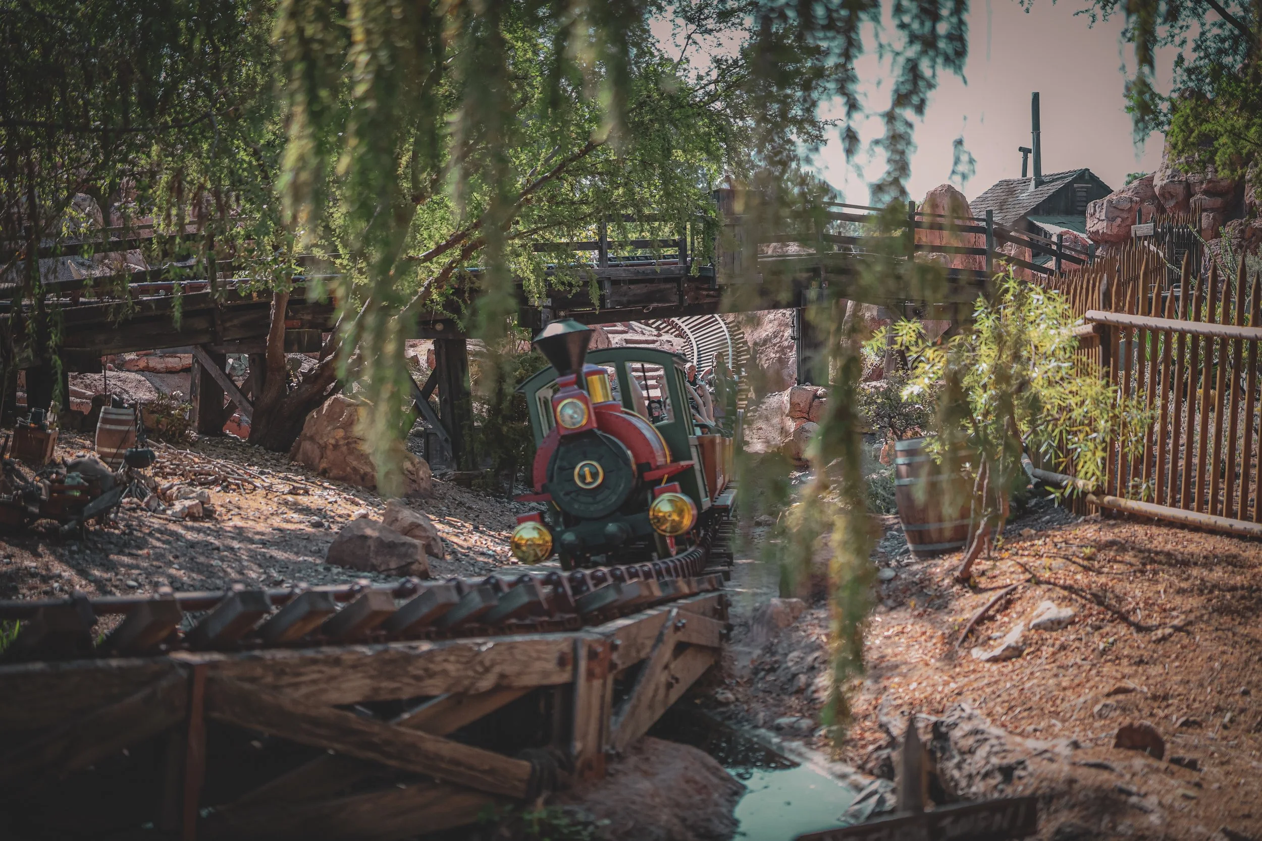 A miniature train on a track within a theme park, passing under a wooden bridge and surrounded by trees, rocks, and wooden fencing.