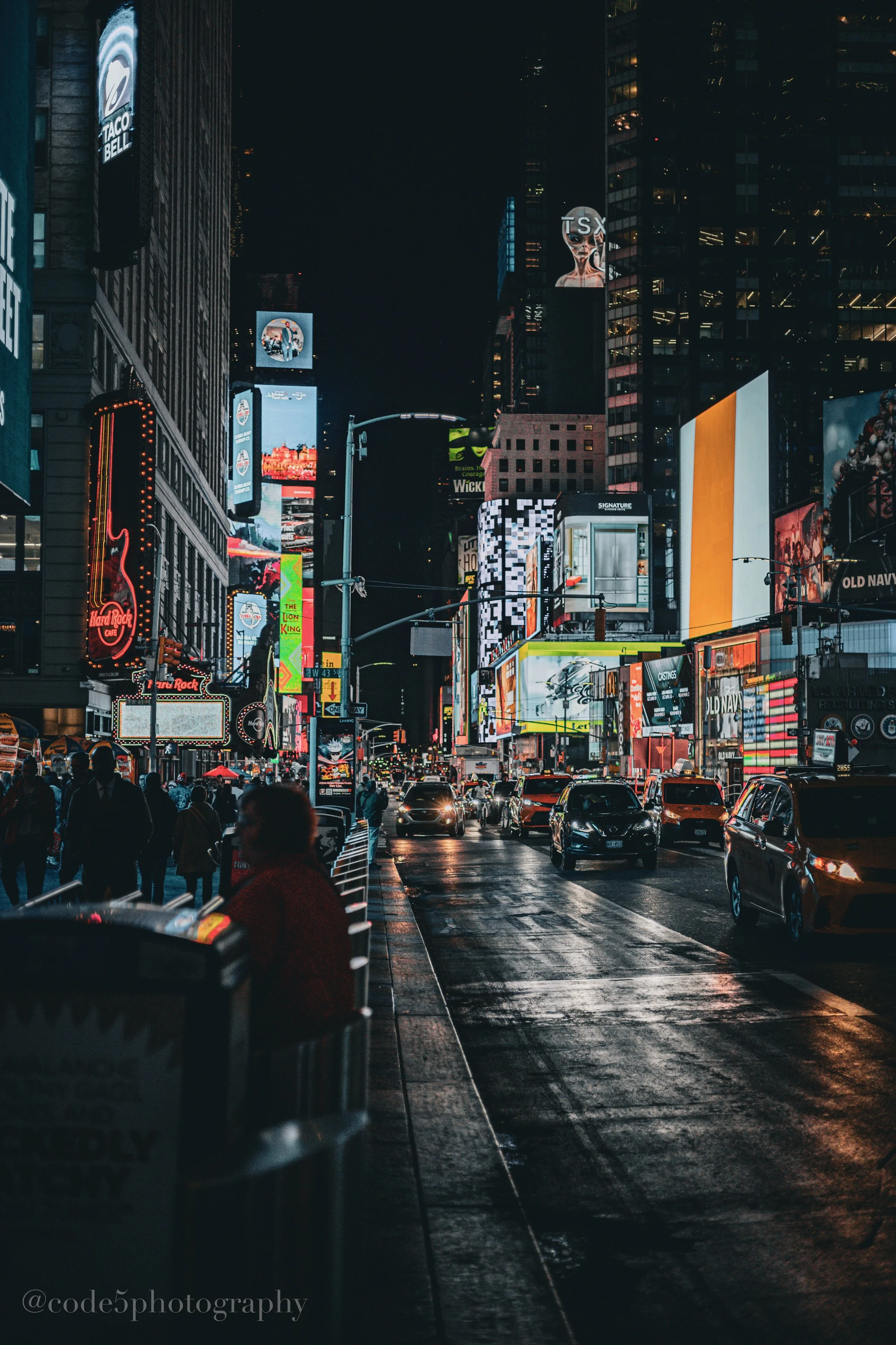 Nighttime scene of a busy city street with cars and people, bright billboards and storefronts, tall buildings, and illuminated advertisements in Times Square, New York City.