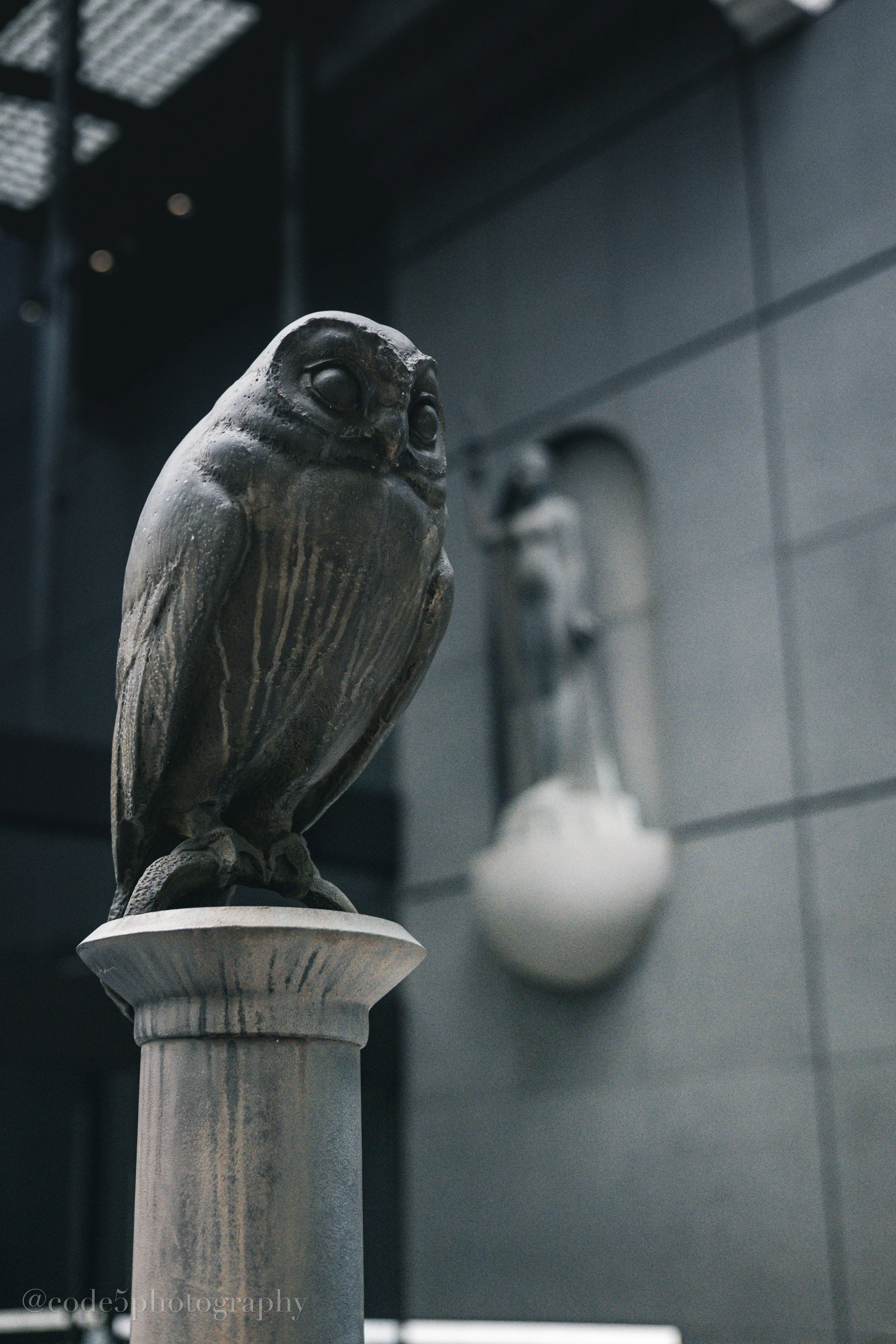 A metal owl sculpture perched on a stone pedestal, with other similar sculptures blurred in the background.