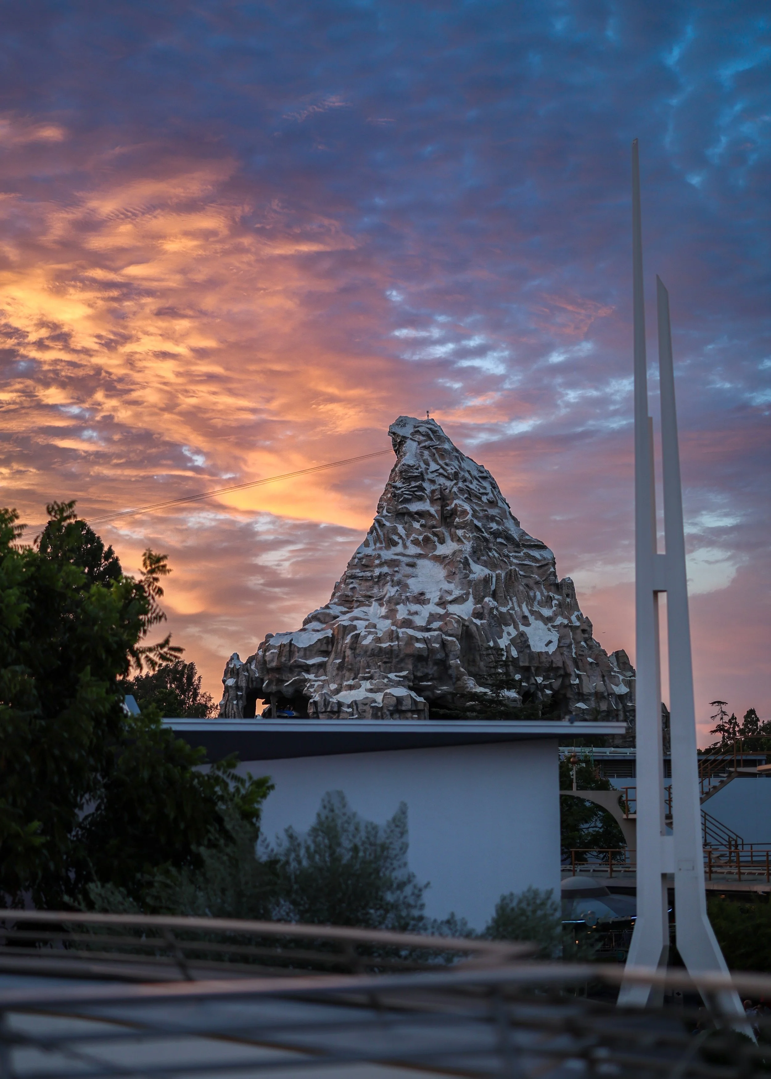 A rocky mountain-like sculpture with snow on top stands against a vibrant sunset sky with pink, purple, and blue clouds. Trees and modern structures are in the foreground.