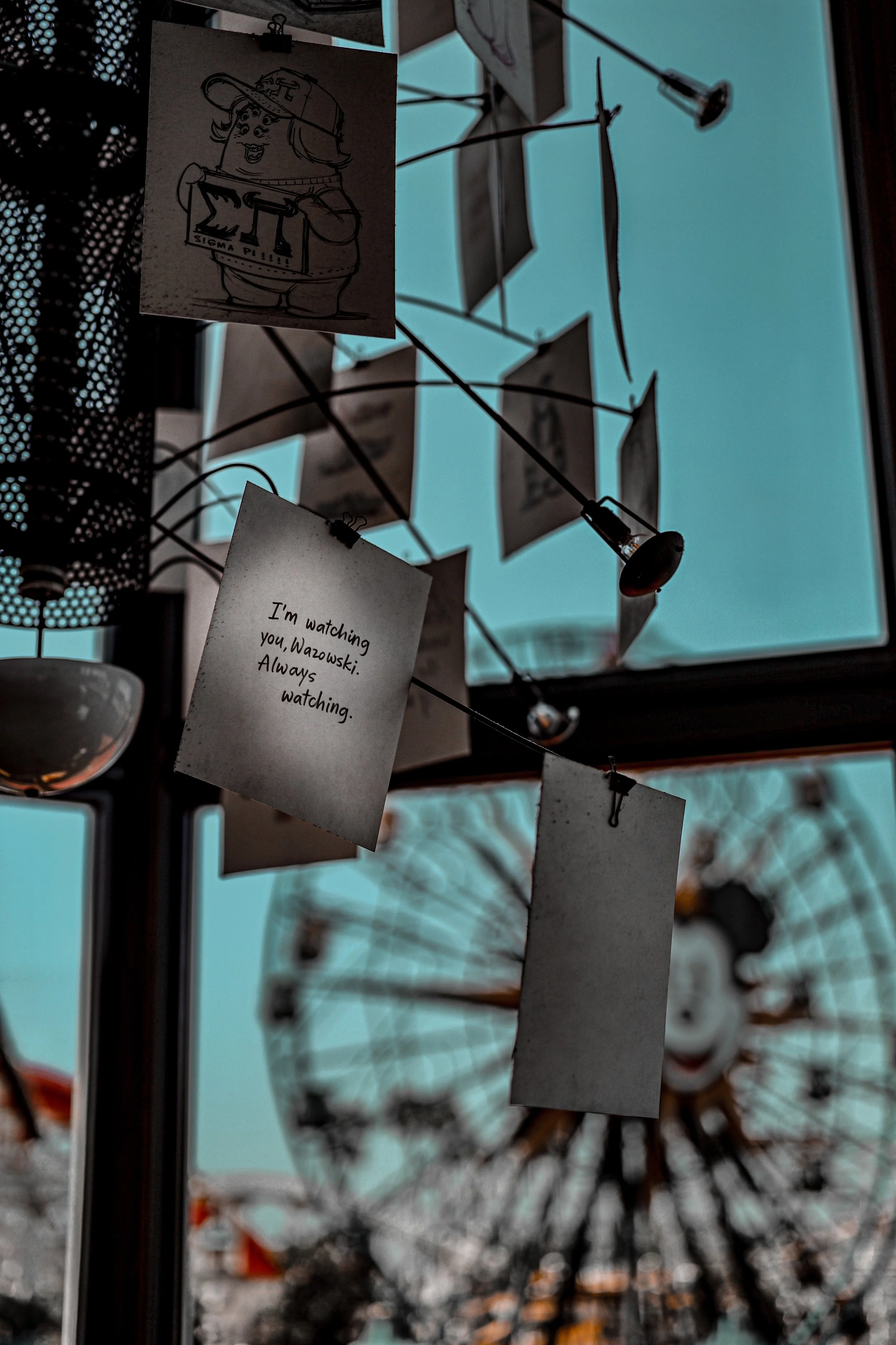 Close-up of a display with hanging notes, including one with a cartoon drawing of a girl in scouting uniform holding a badge, and another note that says, 'I'm watching you, Wazowski: Always watching.' In the background, a diffracted image of a firewo