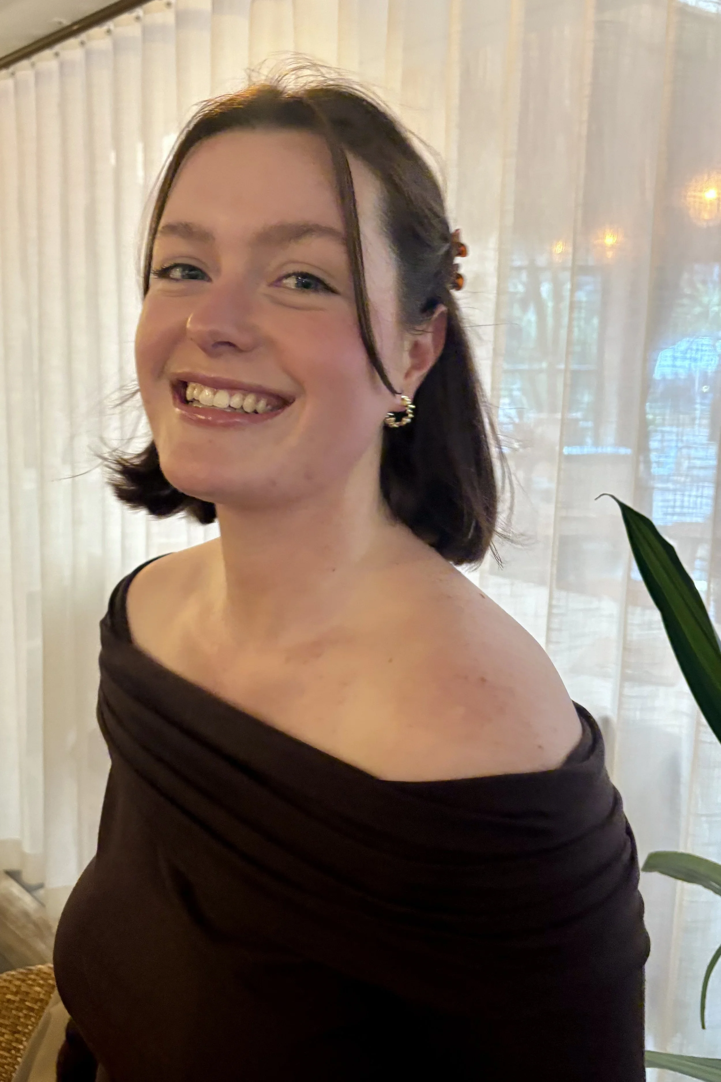 A woman with short dark hair and earrings smiling, wearing an off-the-shoulder black dress, standing in front of sheer white curtains with some greenery visible.