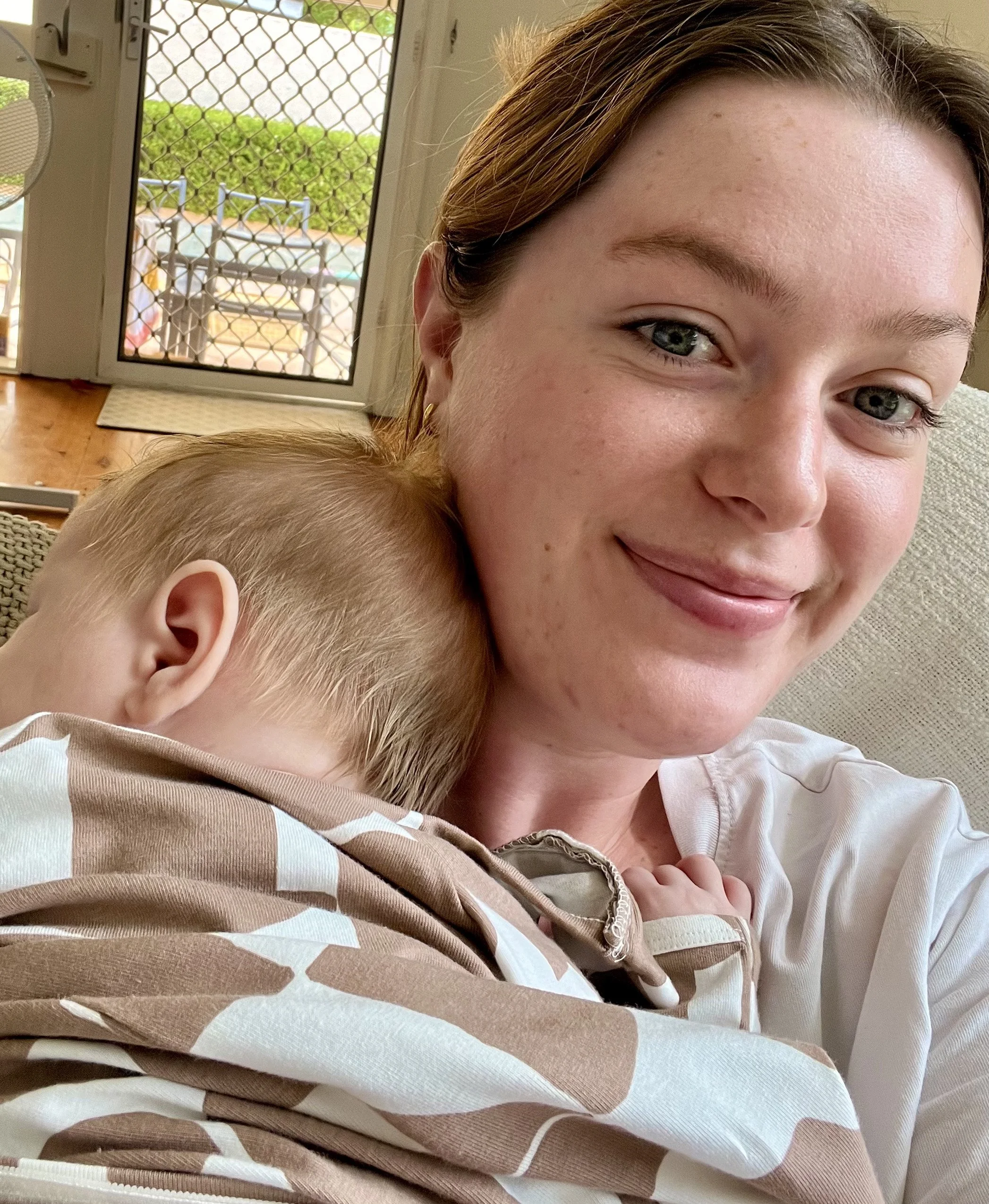 A close-up of a woman's face with her head resting on a child's head, both sitting on a beige couch. The woman has light skin, green eyes, and brown hair. Behind them, there is a sliding glass door with a screen and a view of a backyard with a trampoline and green bushes.