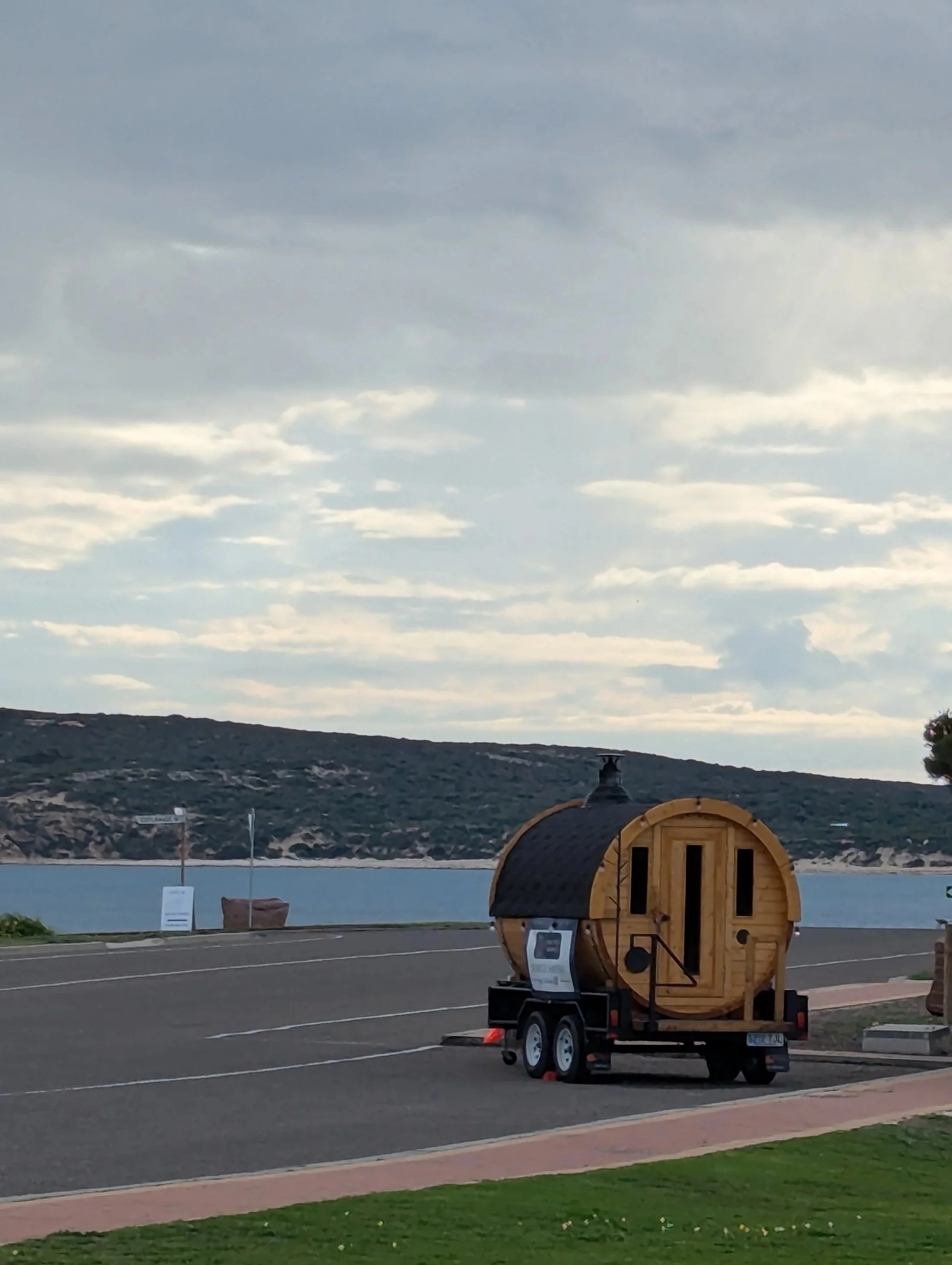 A small wooden sauna on wheels positioned on a paved area near a body of water, with hills and clouds in the background during daytime.