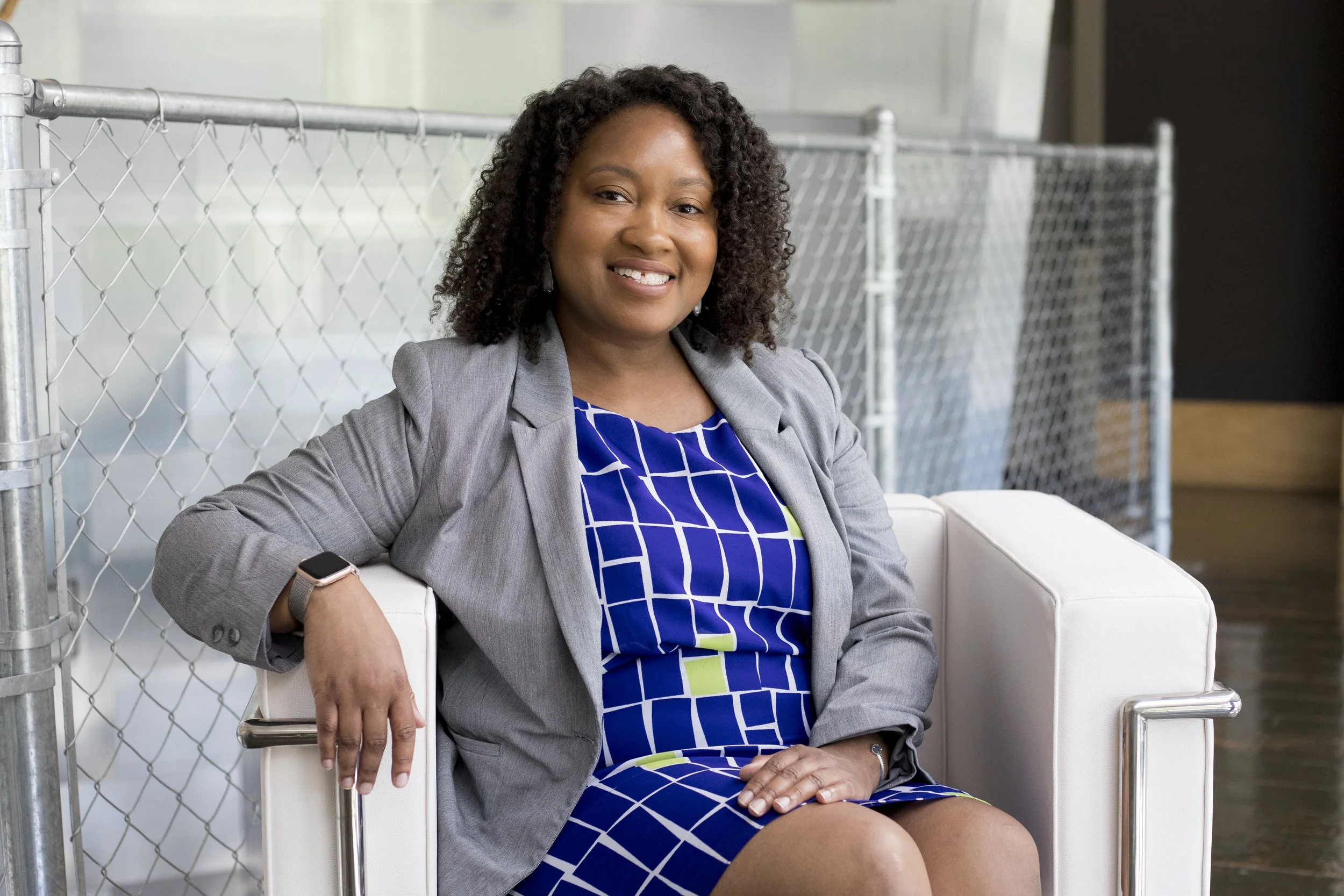 A woman with curly black hair, wearing a gray blazer over a blue and green geometric dress, sitting on a white chair next to a metal fence. She is smiling and has a smartwatch on her left wrist.