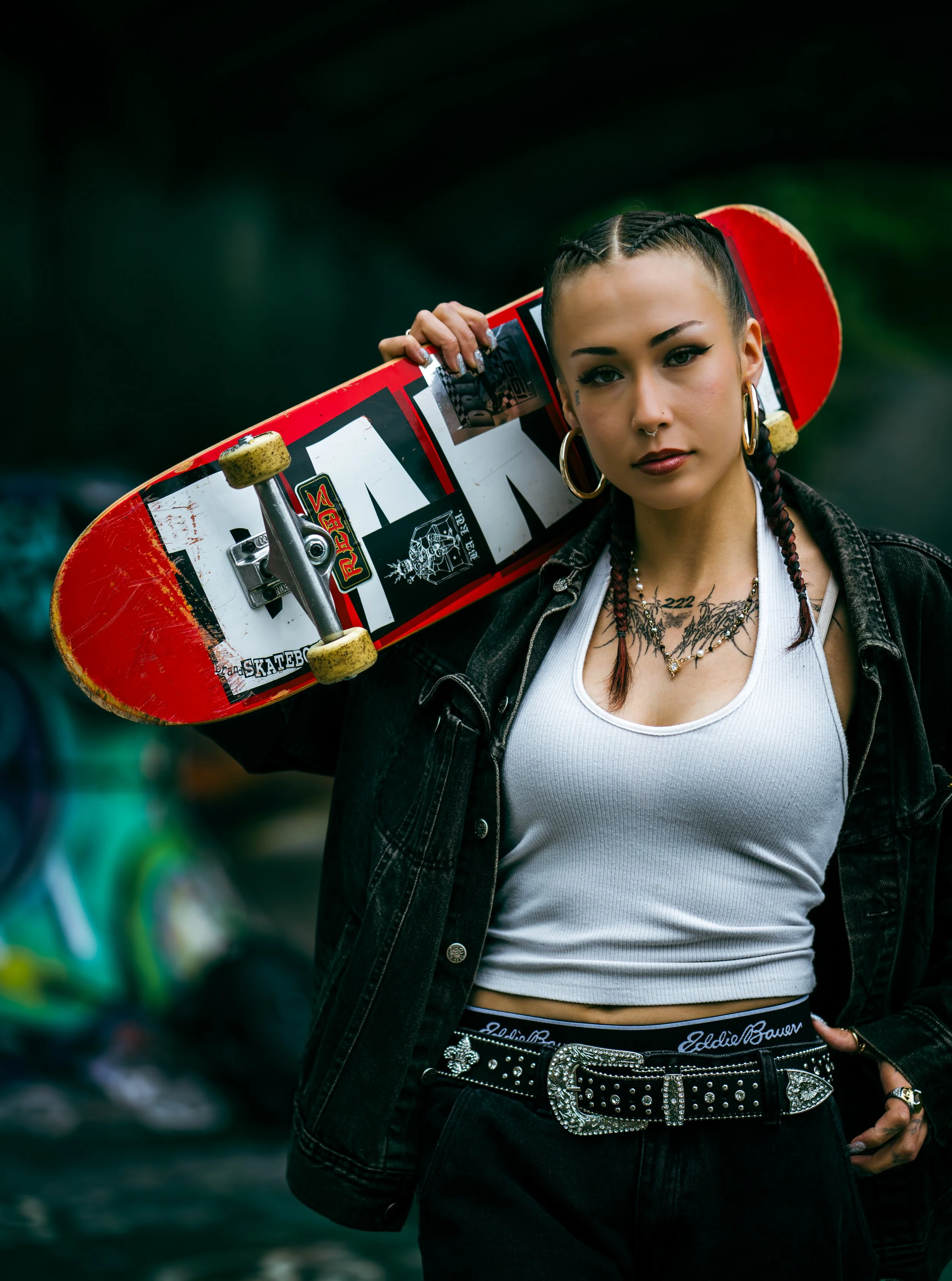 A young woman with braided hair, wearing makeup, jewelry, and a black denim jacket, holding a red skateboard over her shoulder, standing outdoors with a blurred background.