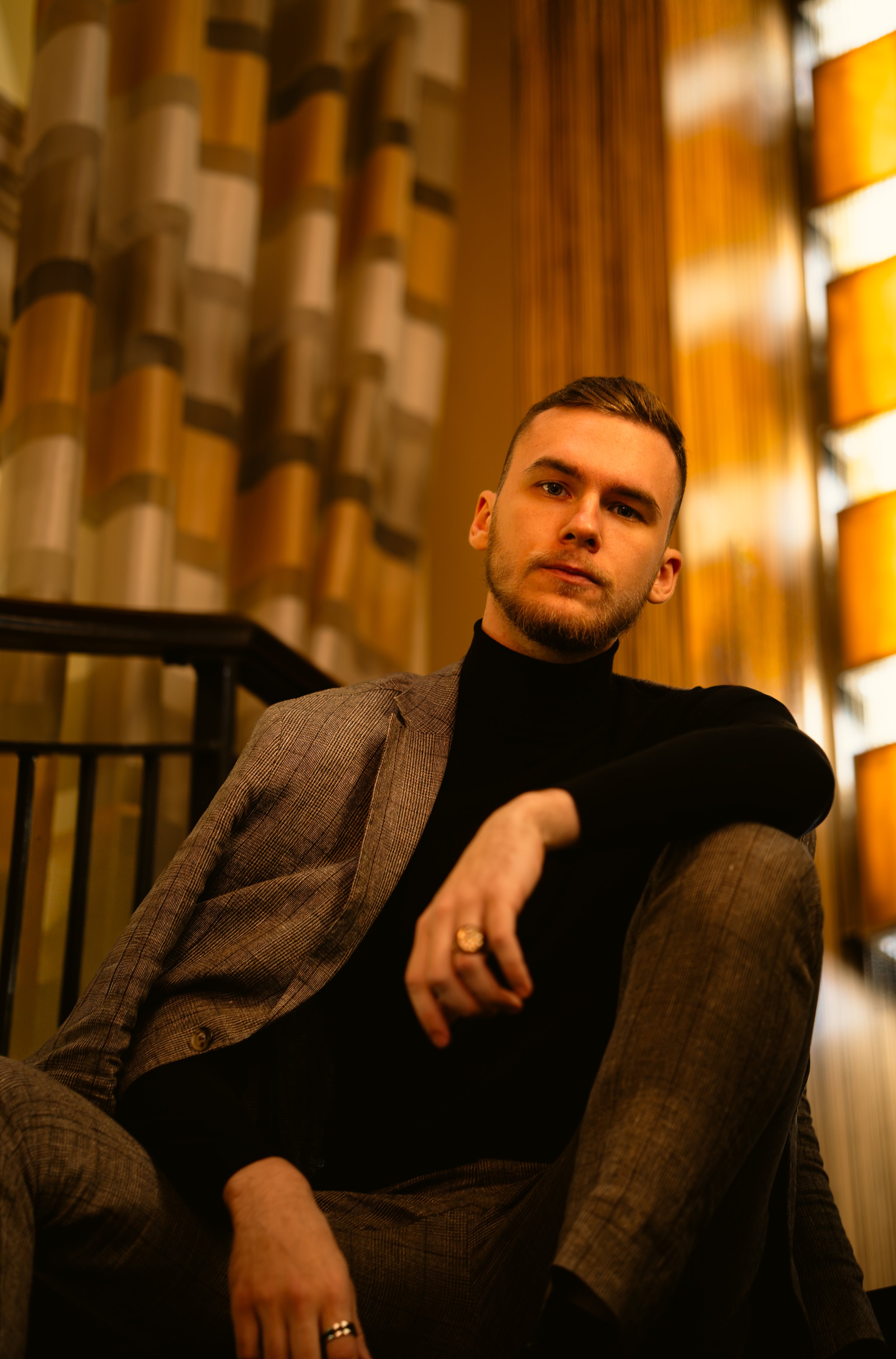 A young man with short brown hair and a beard sitting indoors with a serious expression. He is wearing a dark turtleneck and a brown checkered blazer. The background features warm lighting and wooden wall decor.