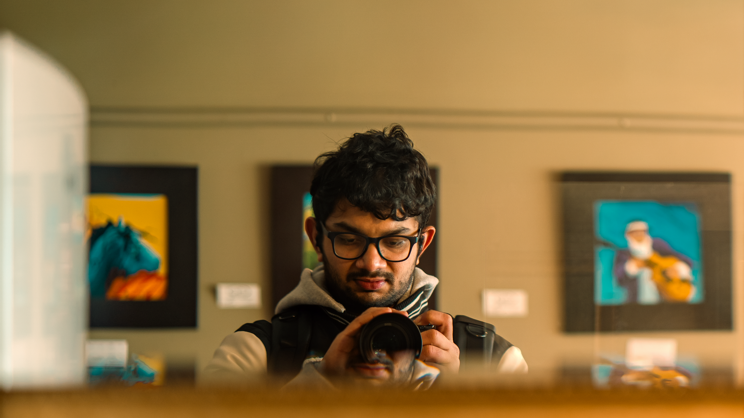 Young man with curly hair and glasses taking a photo with a camera in an art gallery or museum, with colorful artwork on the walls behind him.