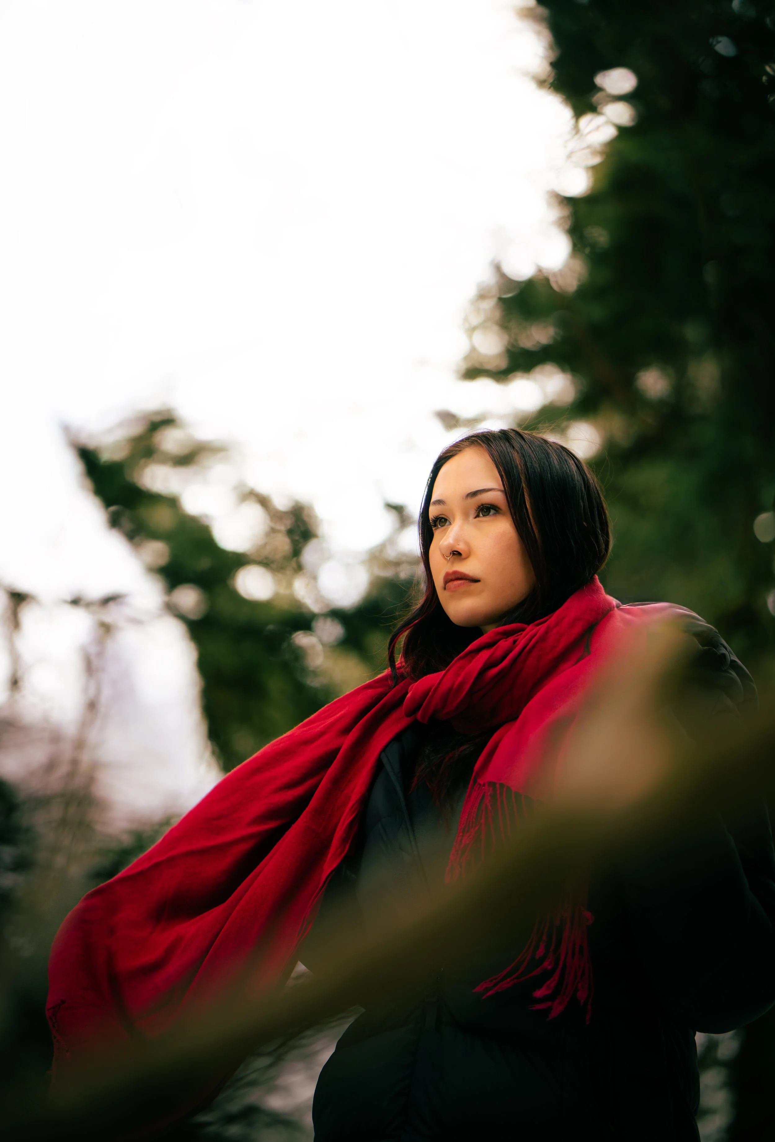 A woman with dark hair, wearing a red scarf and black jacket, standing outdoors with blurred trees in the background.
