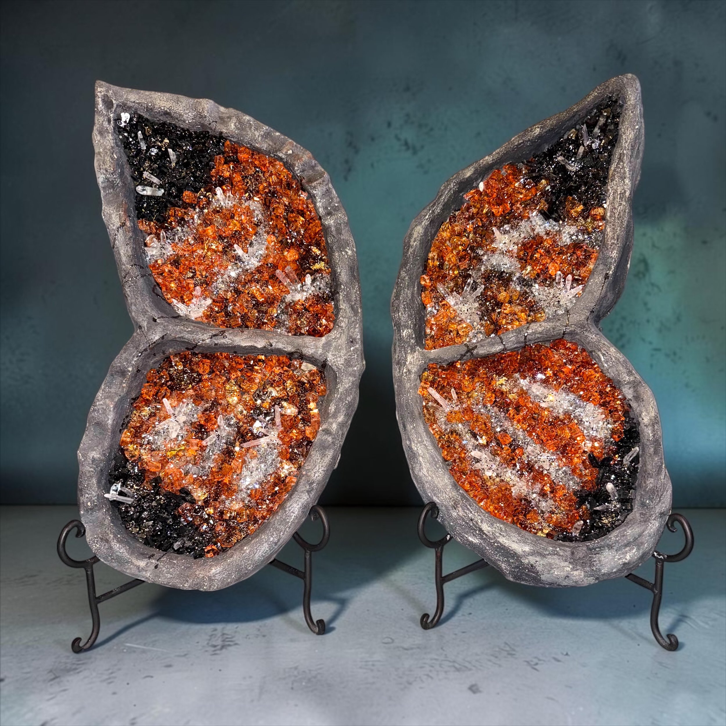 Close-up view of two glass melting vessels filled with colorful crushed minerals. The vessels have a textured surface and are placed on a metal stand with curly legs. A plain, slightly textured background.