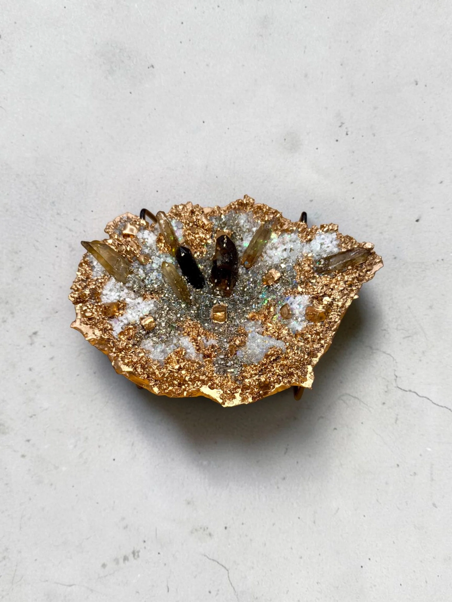 A decorative brooch featuring a gold-tone border and embedded with clear and iridescent crystals, with elongated gold-tone accents radiating outward.