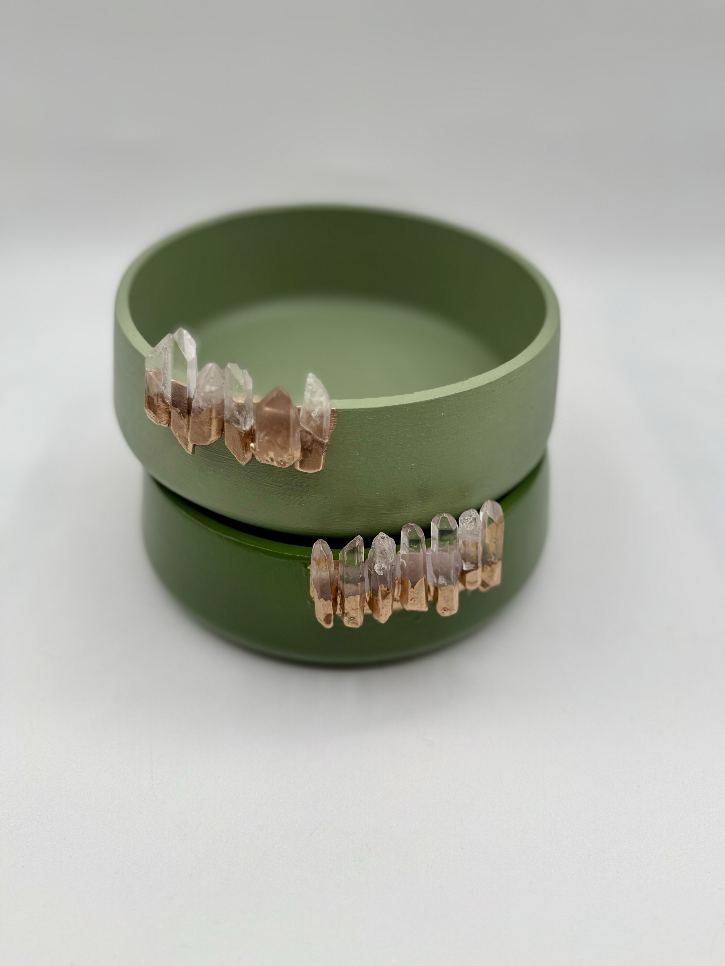 Green circular container with gold and transparent crystal-like beads arranged around its exterior, placed on a white surface.