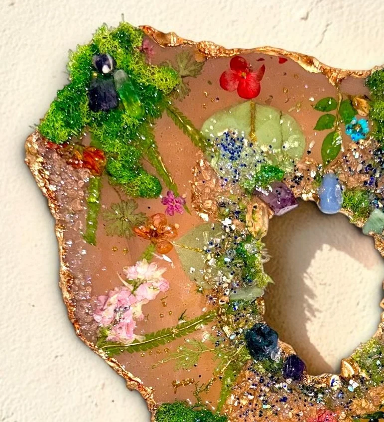 Close-up of a decorative art piece resembling a letter or shield, decorated with various dried flowers, colorful beads, glitter, and textured materials in green, pink, blue, purple, and gold.