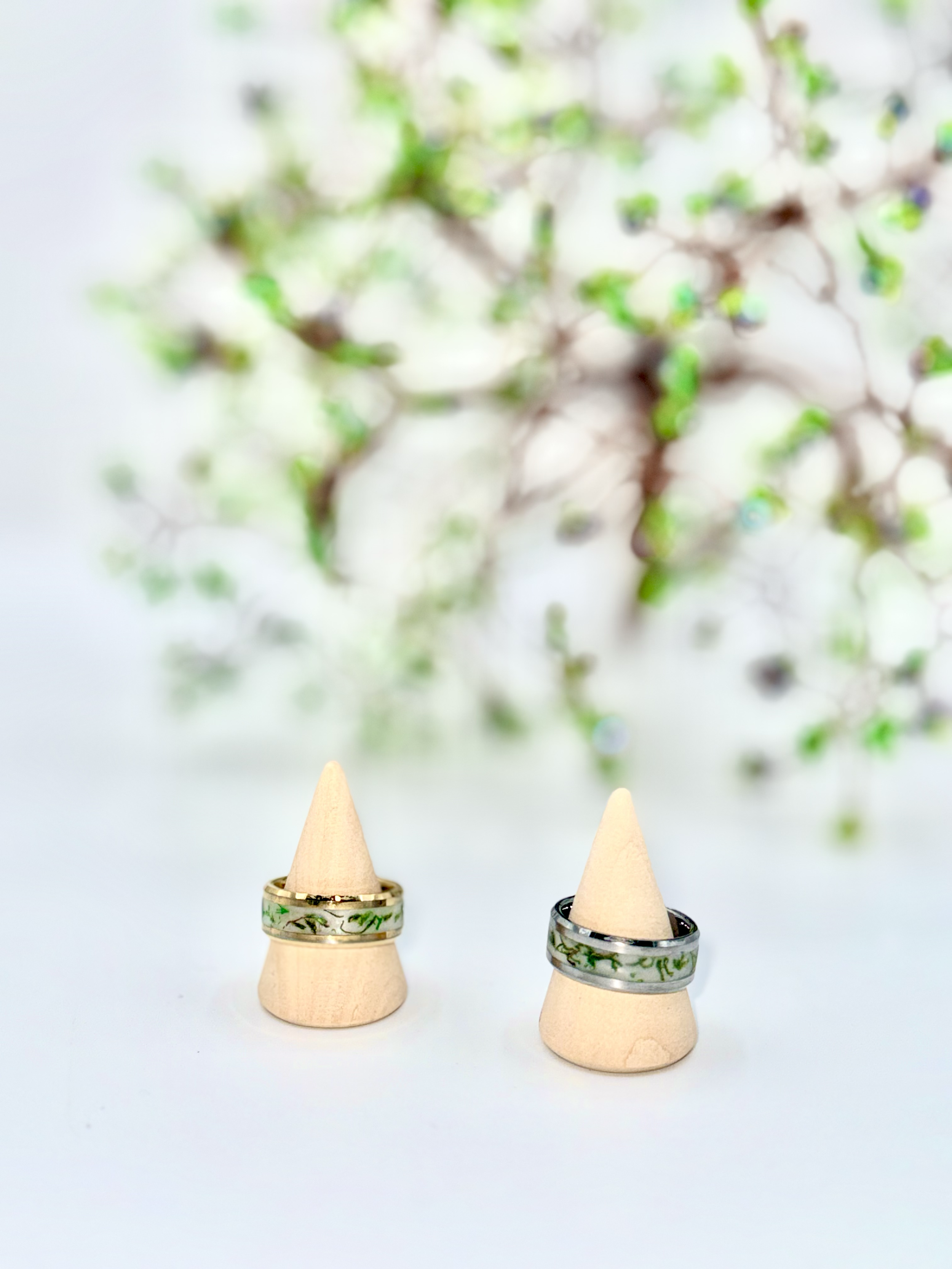 Two wedding rings with green marble pattern are placed on beige ring cones. The background features an out-of-focus display of small green leaves and branches.