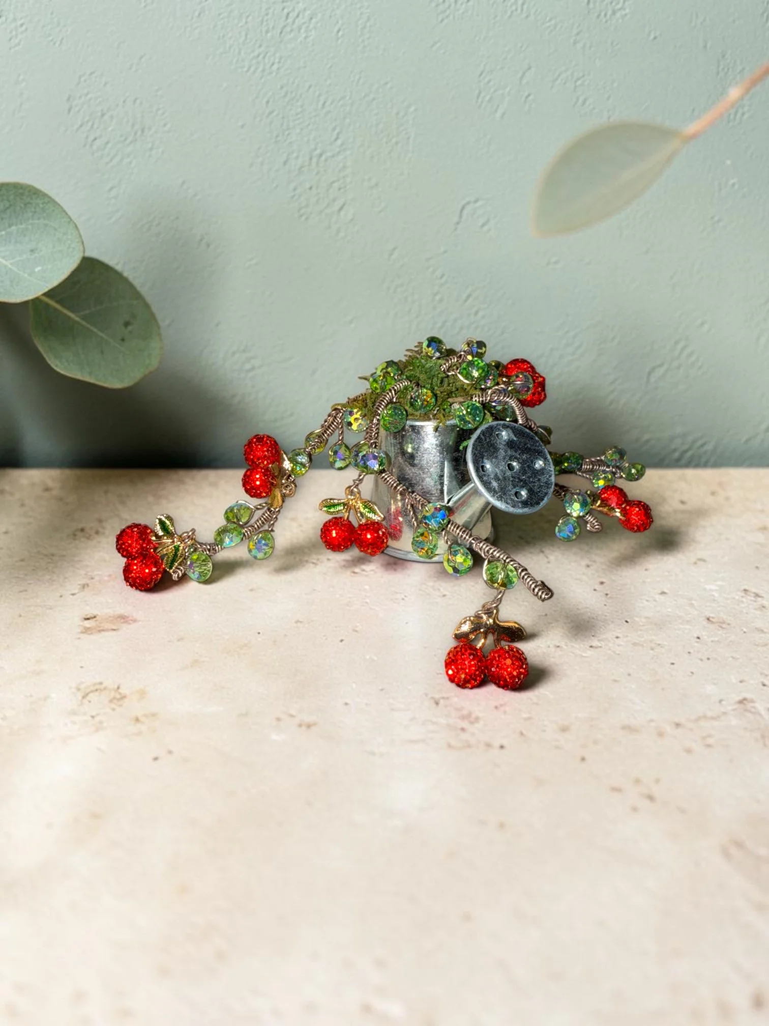 Jewelry with red berry-shaped beads and green leaves, placed on a metal container with silver and colored stones, against a pale green wall and eucalyptus leaves.