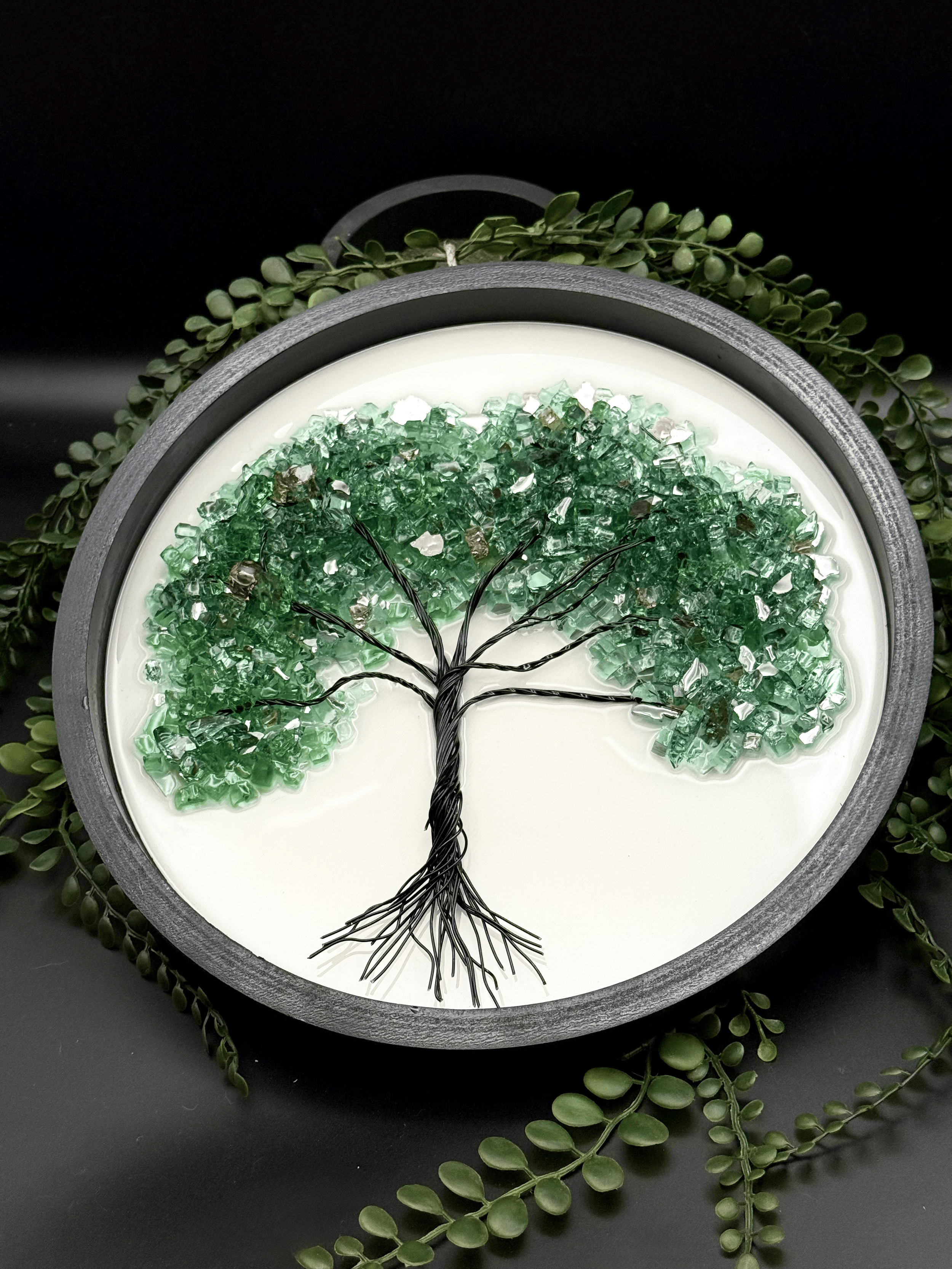 Decorative round wall art featuring a tree made of black wire with green glass pieces for leaves, set against a white background and surrounded by greenery.