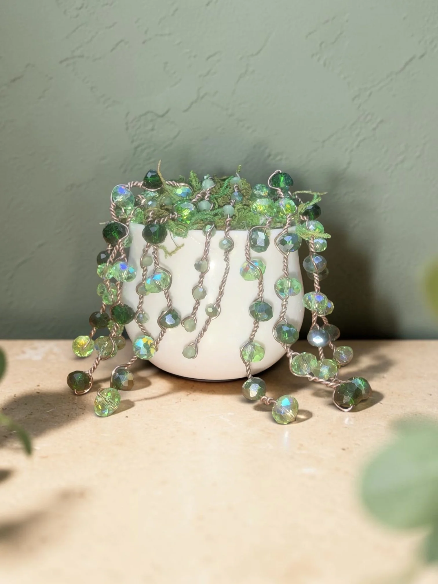 A white ceramic mug filled with green moss and adorned with dangling iridescent green and clear beads on rose gold wires.