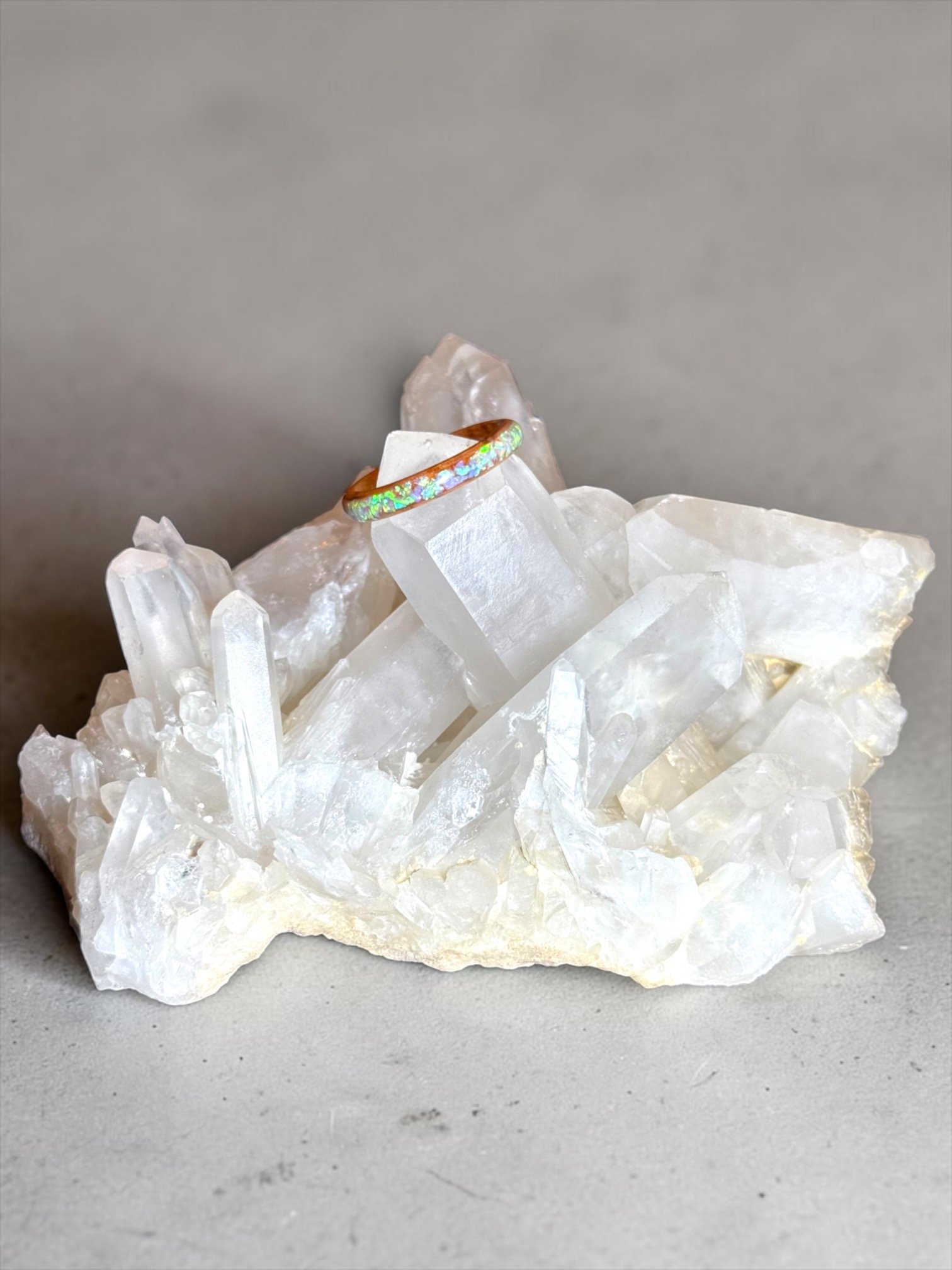 A rainbow-colored ring resting on a large, clear quartz crystal cluster, with a gray background.