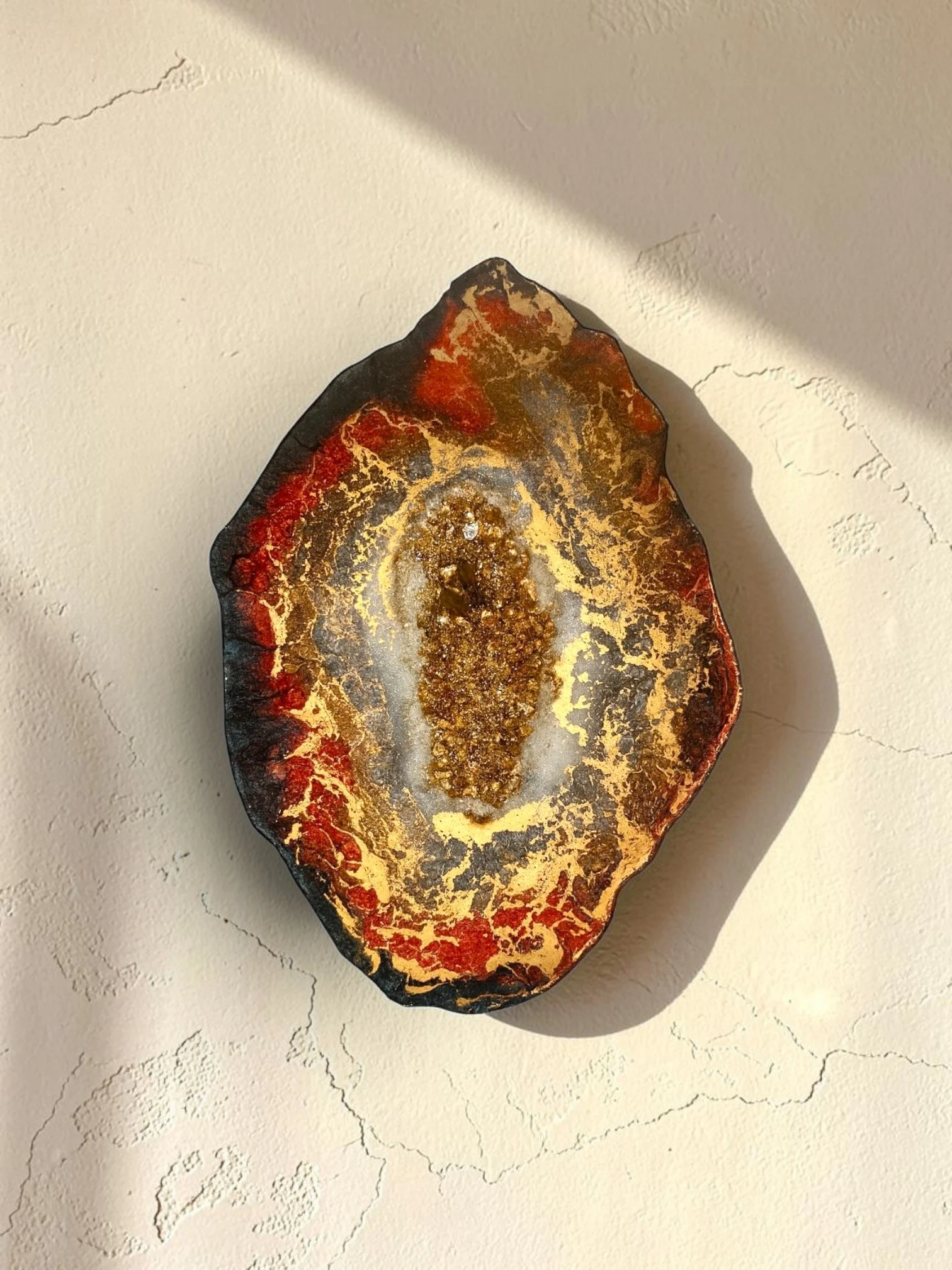 A polished, irregularly shaped mineral or gemstone with red, black, and gold colors, resting on a white textured surface with faint cracked pattern.