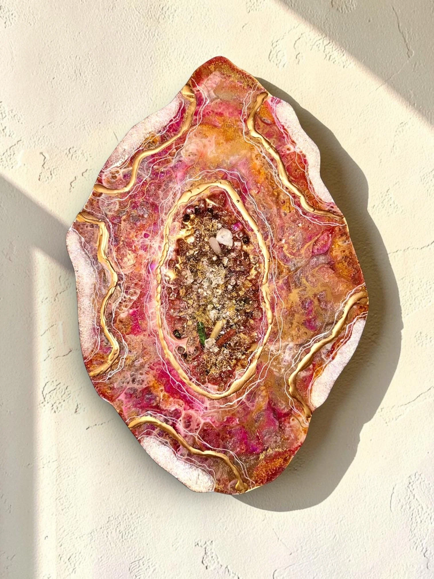 A colorful, polished geode with pink, gold, and brown hues, showing intricate crystalline patterns and a reflective surface hanging on a white textured wall, casting a shadow.