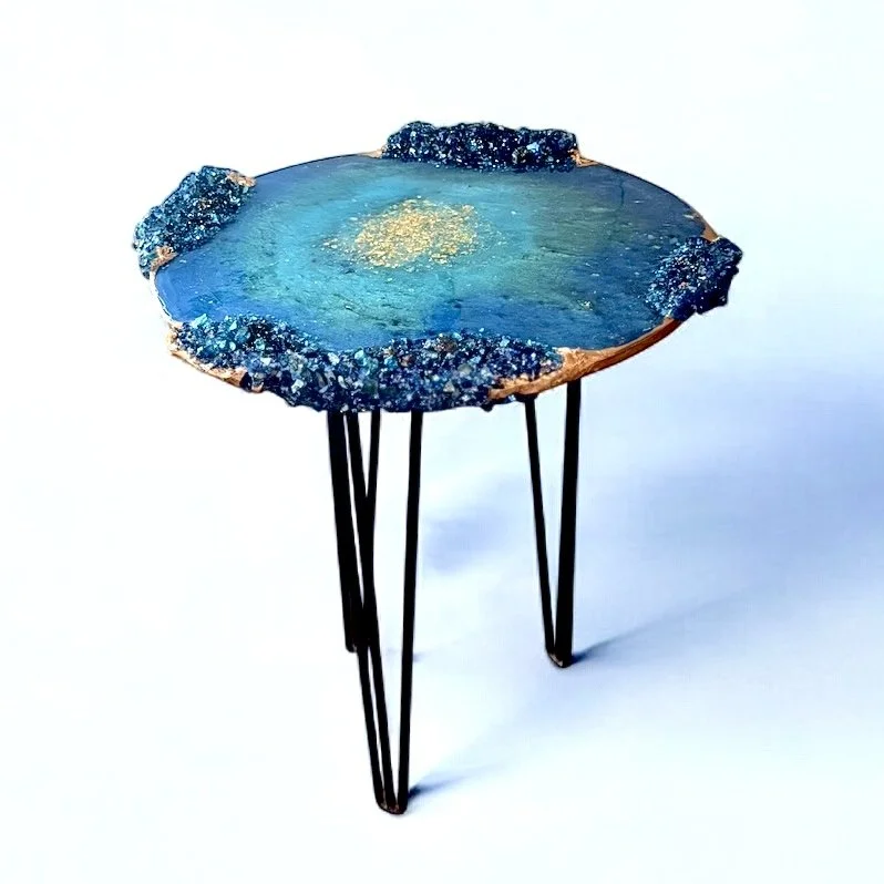 A small side table with a round, polished wood top and textured blue resin edges, supported by three black metal hairpin legs.