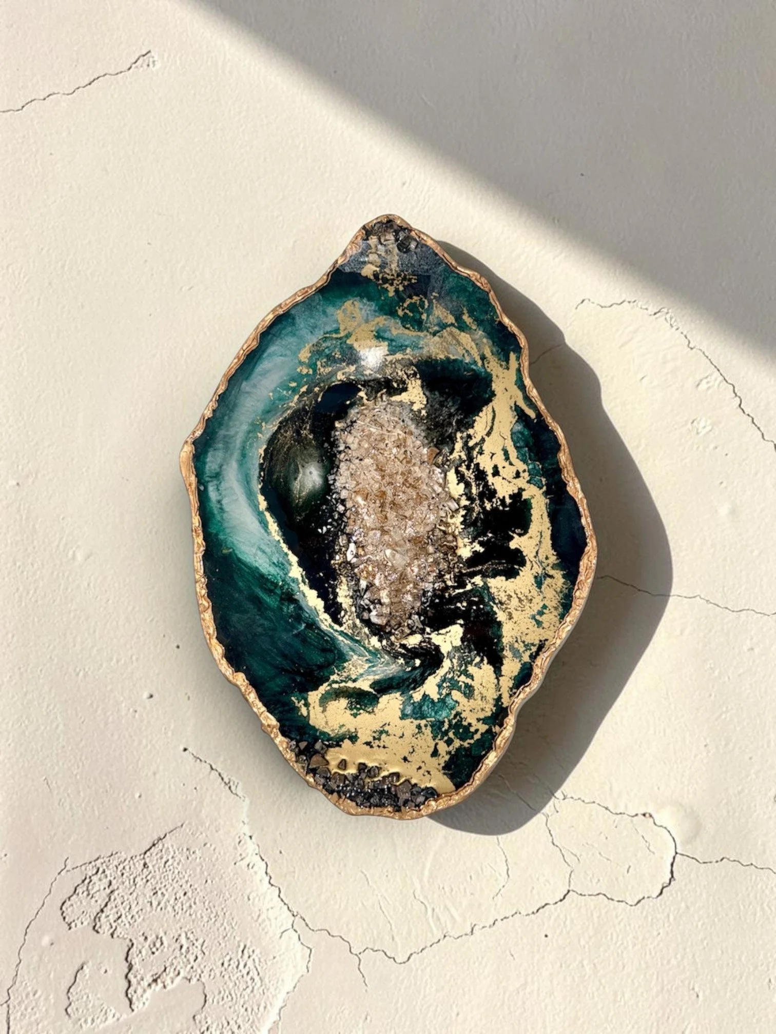 A polished geode with an irregular edge, showing a dark green and black interior with gold accents, placed on a white textured surface with visible cracks.